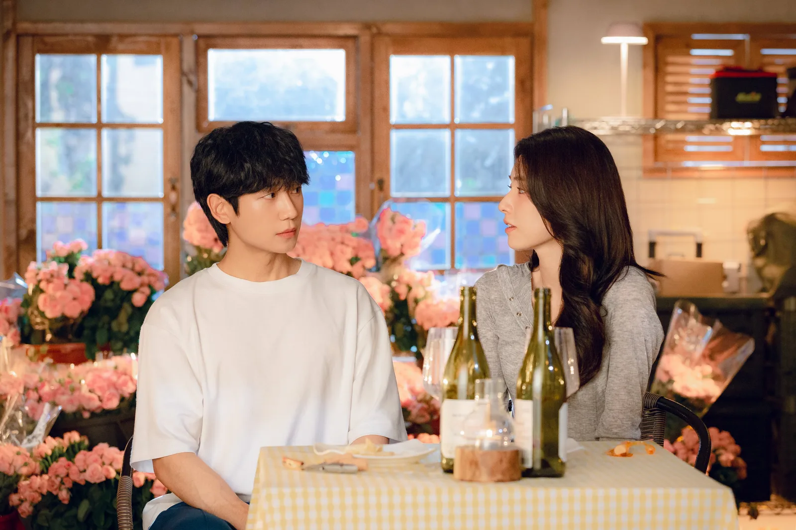 This image captures a candid moment from the Netflix romantic comedy series Our Sticky Love (2026), starring Jung Hae-in as small-town boxing coach Jang Tae-ha and Ha Young as the amnesiac prosecutor Go Eun-sae. In this scene, the two leads are seated opposite each other at a cozy dinner table covered in a yellow-and-white checkered tablecloth, surrounded by a romantic yet slightly overwhelming arrangement of abundant pink rose bouquets in the background. Tae-ha, dressed casually in an oversized white t-shirt, gazes toward Eun-sae with a mixture of quiet attentiveness and hidden anxiety, while Eun-sae, wearing a simple gray button-up shirt, looks back at him with an expression of subtle suspicion and curiosity as she tries to parse through her forgotten past. Between them on the table sit two green glass wine bottles, a rustic candle holder, and a small white plate, framing their tense, bickering dynamic in a warm, warmly lit interior that highlights the show's signature mixture of small-town romance and undercover secrets. 