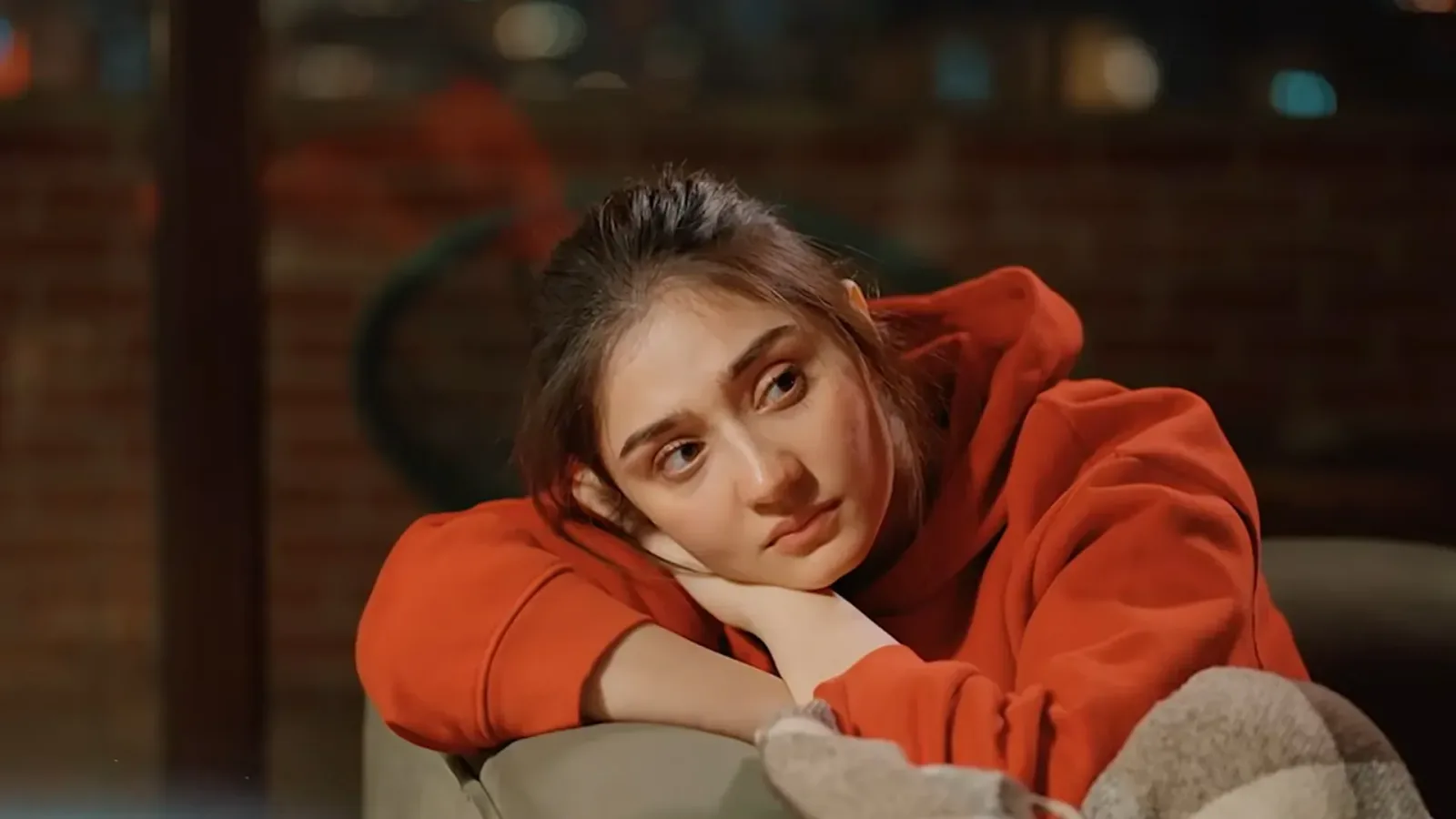 A close-up shot capturing actress Dur-e-Fishan Saleem resting her chin on her hand while leaning over the back of a muted beige sofa couch. She is wearing a vibrant, oversized bright orange hoodie with the hood pulled back, slightly gathering around her neck, and her dark brown hair is pulled back into a casual, soft bun with delicate loose strands framing her face. Her expression is deeply pensive, melancholy, and contemplative, with her dark brown eyes looking off into the distance, lost in thought. The background is softly blurred with a warm, dim low-light ambiance, showcasing a rustic brick wall texture and faint indoor lighting bokeh that emphasizes her isolated, reflective, and sorrowful mood.