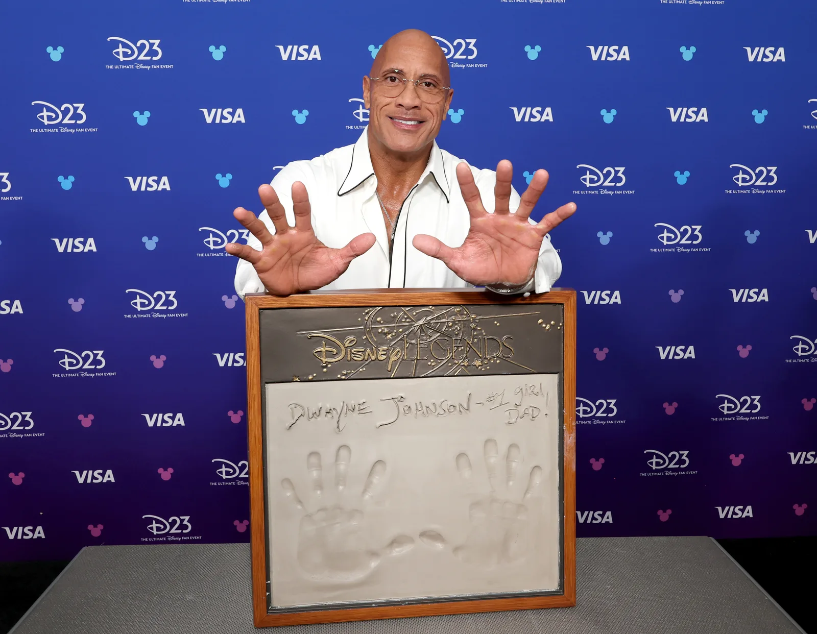 Dwayne Johnson honored by 'Jungle Cruise' costar Emily Blunt. An imposing, muscular man with a shaved head and a warm smile stands behind a rectangular plaque, holding up both of his hands with fingers spread wide to match the handprints cast into the cement below. He is wearing a crisp, open-collared white button-down shirt. The framed cement plaque features his signature, "Dwayne Johnson," inscribed above the deeply indented handprints. The background is a solid royal blue step-and-repeat wall covered in a repeating pattern of white logos including "D23" and "VISA," creating a promotional or ceremonial atmosphere.