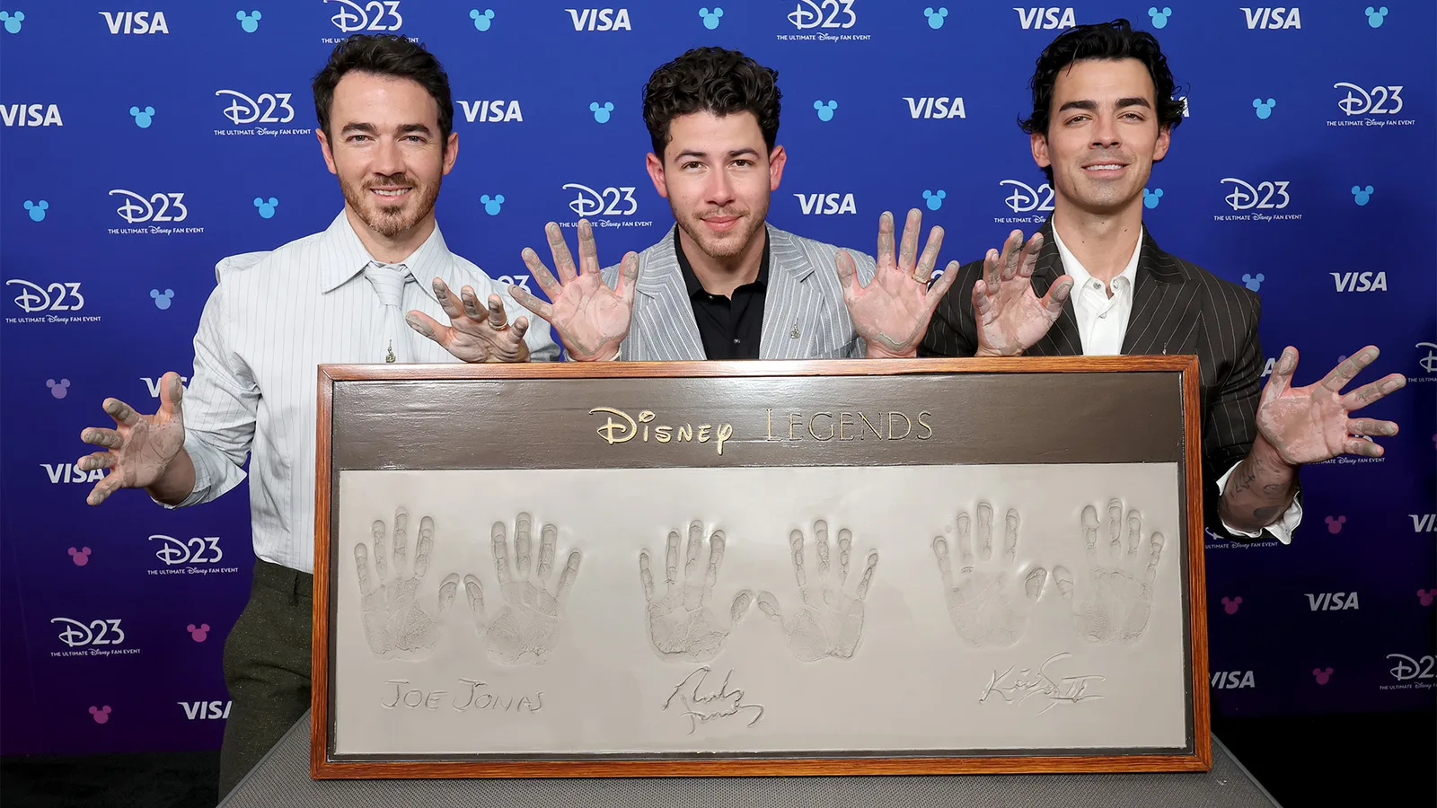 Jonas Brothers honored as Disney Legends by Vanessa Hudgens - An image featuring Kevin Jonas, Nick Jonas, and Joe Jonas standing behind a large framed cement plaque containing their handprints and signatures. The trio is posing with their hands raised, showing cement residue on their fingers, against a blue Disney D23 and Visa branded backdrop.