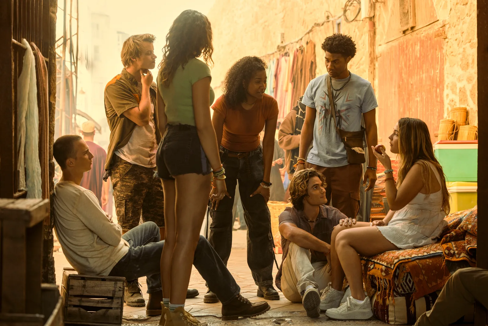 A group of seven young actors from the television series Outer Banks are gathered together in an alleyway marketplace with a warm, golden sunset glow. In the foreground, a girl with her back turned wears a green top and dark shorts, while to her left, a boy sits on a wooden crate leaning against a wall. Two girls and three boys stand and sit in a tight circle in the center, engaged in conversation. The background features stone walls, hanging textiles, and market stalls, creating a warm, dusty atmosphere.