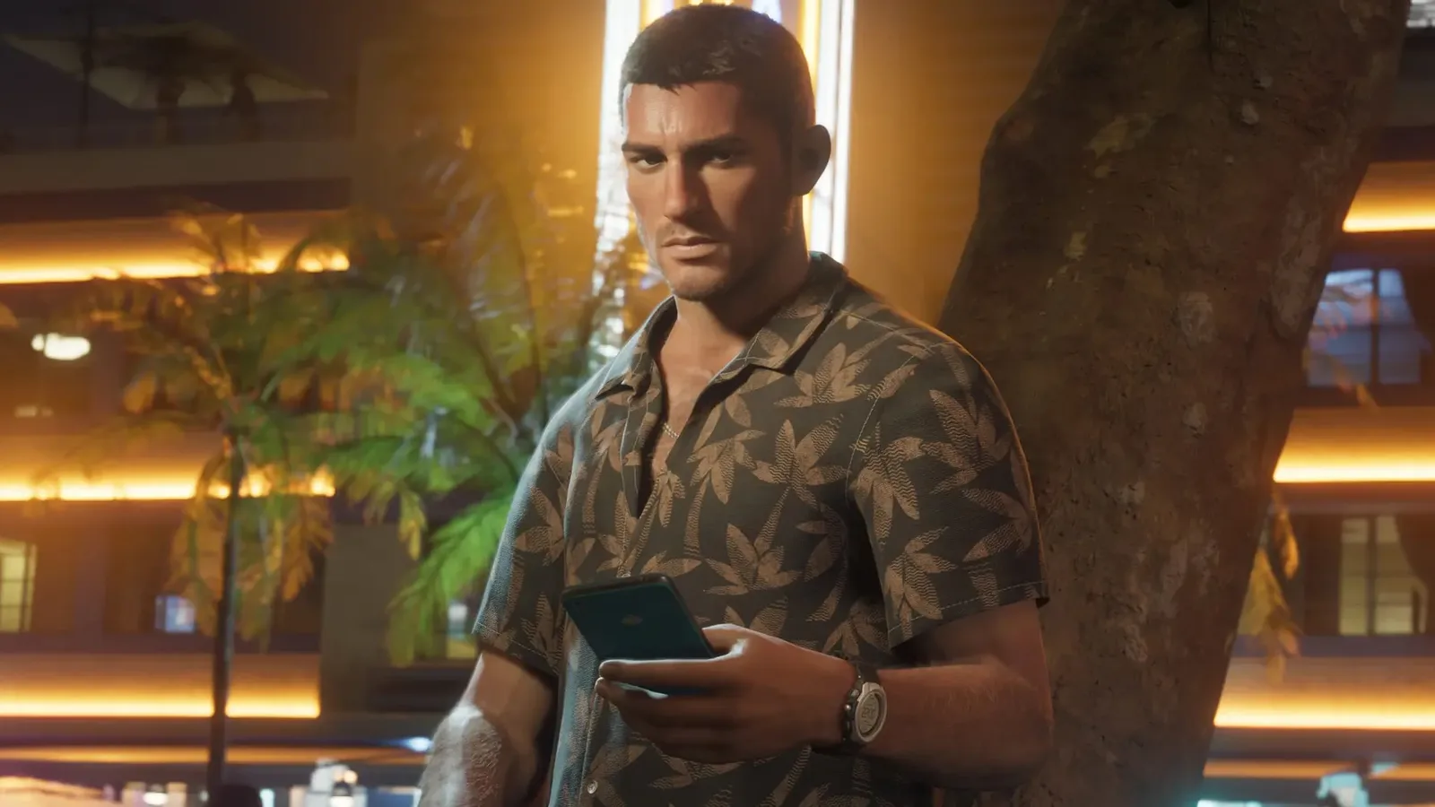 An attractive young man with short dark hair, wearing a brown Hawaiian shirt patterned with beige palm leaves, stands outdoors at night holding a smartphone. He leans slightly toward a large tree trunk on his right, while a warmly illuminated modern building and palm trees create a vibrant, glowing backdrop behind him. Looking directly forward with a serious, intense expression, he holds the phone in both hands, revealing a wristwatch on his left arm.