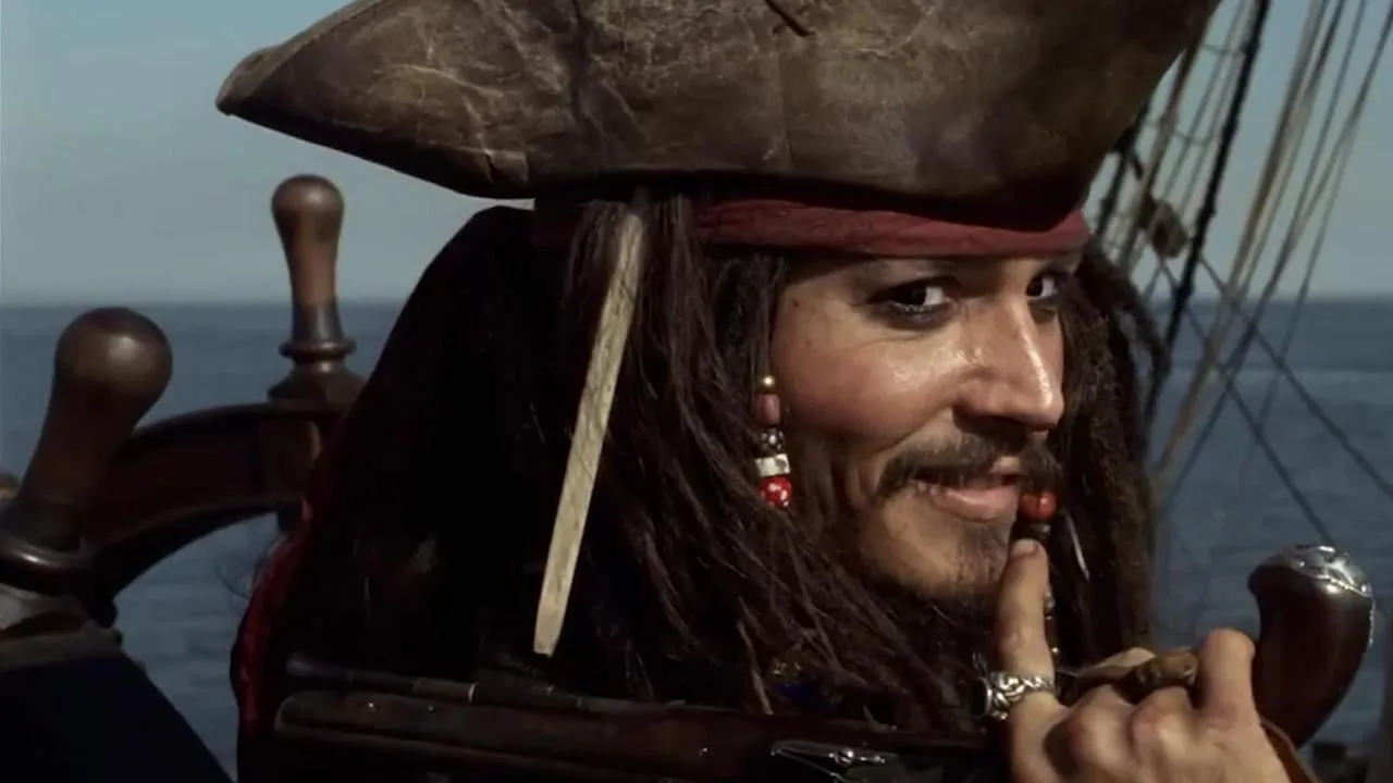A close-up shot captures Captain Jack Sparrow standing at the helm of a ship, looking over his shoulder toward the camera with a subtle, mischievous smirk. He wears his signature weathered brown tricorn hat over a red bandanna, with his dark, dreaded hair decorated with hanging beads and a distinctive long bone ornament. His face features dark eyeliner, a thin mustache, and a braided goatee, while one hand rests near his chin, showing off large silver rings and a flintlock pistol gripped nearby against the backdrop of the open blue ocean and ship rigging.