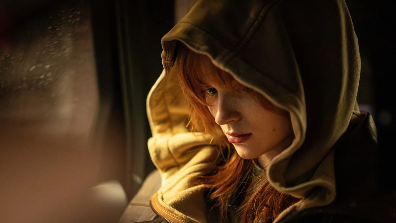 A cinematic close-up shot captures American actress Sadie Sink in her role as Jean Grey from Spider-Man: Brand New Day. She sits inside a dark vehicle, looking downward with a pensive, intense expression. Her signature red hair frames her face, partially obscured by the large, oversized hood of a beige or olive-toned zip-up hoodie. She wears a dark leather jacket over the hoodie. Warm, low-key lighting from the front illuminates her facial features and the inner texture of the hood, contrasting sharply against the shadowy background. Through the window to her left, soft rain streaks across the glass pane, adding to the moody and secretive atmosphere of the scene.
