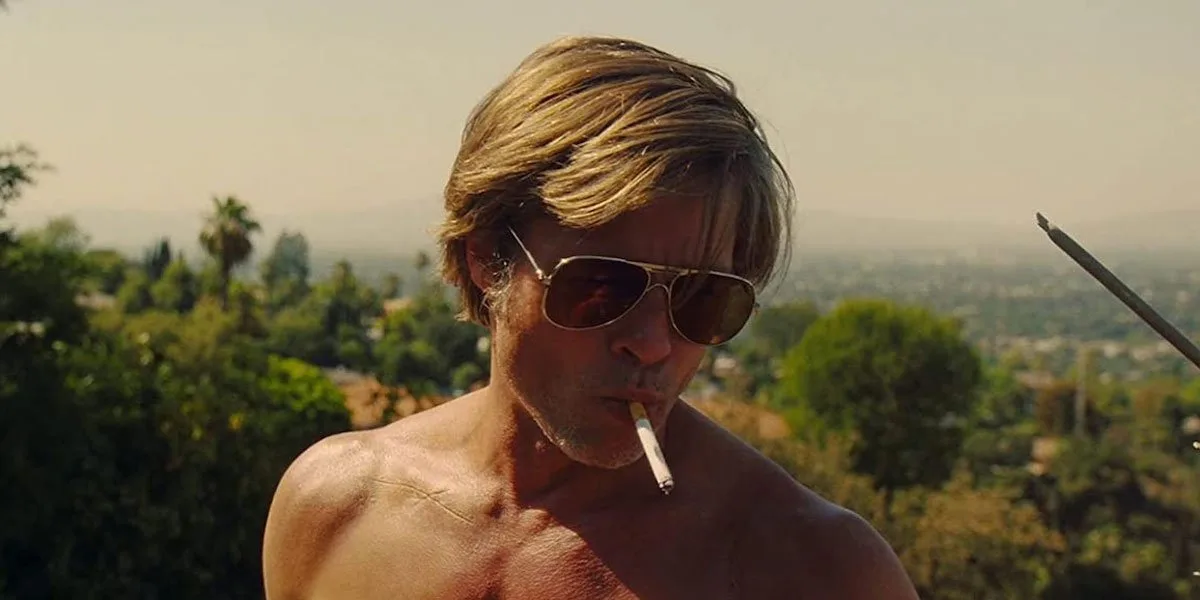 does brad pitt smoke cigarettes in real life Was Able To Go Incognito And  Help Those Need Thanks To Mask-Wearing Brad Pitt's Smoking Habit Worries –  A medium close-up shot from the film Once Upon a Time... in Hollywood captures a shirtless, muscular Brad Pitt portraying the character Cliff Booth against a hazy, sun-drenched landscape of Los Angeles hills. His golden-blonde hair is neatly parted and styled in a classic late-1960s fashion, gently catching the warm afternoon sunlight. He wears gold-rimmed aviator sunglasses with dark, reflective lenses that obscure his eyes, adding to his rugged and detached expression. A white cigarette hangs casually from his lips as he prepares to light it while standing outdoors on a rooftop. The background features a soft-focus panorama of lush green trees, rolling hills, and a clear, bright sky, emphasizing the hot, dry climate of Southern California. A metal television antenna pole enters the right side of the frame, hinting at the rooftop setting where the character is working. The overall color palette is saturated with warm yellow and golden tones, characteristic of Quentin Tarantino's vintage cinematic style.