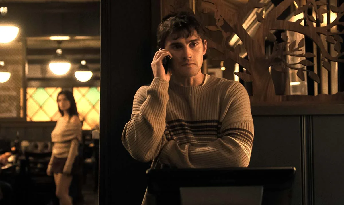 A dimly lit restaurant interior from the film Obsession, where Bear (played by Michael Johnston) stands in the foreground behind a terminal counter, looking tense and anxious while talking on a smartphone held to his ear. He is dressed in a beige, horizontally striped crewneck sweater. In the blurred background to the left, his childhood friend Nikki (played by Inde Navarrette) stands near the dining booths, wearing an off-the-shoulder light sweater and dark miniskirt, looking toward him. An ornate, laser-cut wooden divider shaped like tree branches framing the scene on the right provides a subtle artistic detail to the establishment's decor.