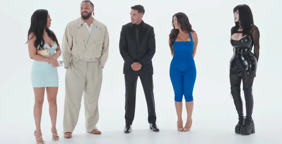 A full studio shot captures five individuals standing side-by-side against a stark white backdrop, each wearing vastly contrasting styles of clothing. On the far left, a woman wearing a light blue sleeveless mini dress and strappy high heels turns her head toward the man next to her while holding a small clutch. The man beside her wears a voluminous, oversized tan trench coat paired with matching wide-leg trousers and brown dress shoes. In the center stands a man clad in a tailored black suit with a black dress shirt and dark tie, looking toward the woman in blue on his left. That woman wears a vibrant, fitted royal blue sleeveless jumpsuit that cuts off below the knee, paired with nude heels. On the far right stands a woman styled in a dramatic gothic aesthetic, wearing a black latex or leather corset top, a matching mini skirt, sheer black sleeves, and thigh-high black platform boots.