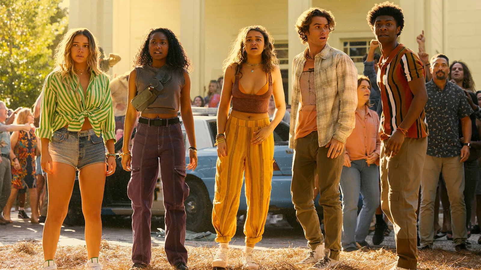 A production still from Netflix's adventure drama series Outer Banks, capturing five main characters—Sarah Cameron, Cleo, Kiara Carrera, John B Routledge, and Pope Heyward—standing in a row out of doors during a warm, sunlit afternoon. From left to right, Sarah wears denim shorts and a tied, green-and-white vertically striped button-down shirt; Cleo is dressed in purple cargo pants and a gray sleeveless crop top with a prominent tactical cross-body chest bag; Kiara wears flowing, orange-and-yellow vertically striped pants paired with a dark orange crop top; John B stands in a white plaid open-button shirt layered over a light orange tank top and khaki pants; and Pope stands on the far right wearing a vertically striped, short-sleeve polo shirt with tan trousers. The group exhibits expressions of intense shock, confusion, and concern as they gaze off-camera to the right. Behind them, a white and blue local police vehicle is visibly parked amidst a scattered, blurred crowd of onlookers under the golden-hued sunlight.
