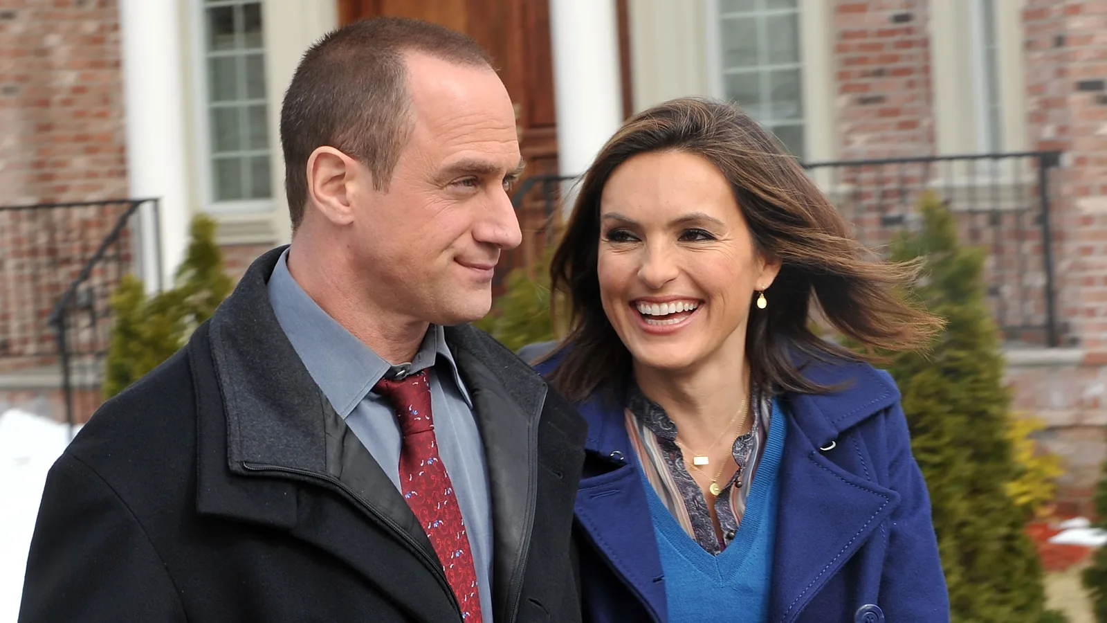 An intimate and candid moment captures actors Christopher Meloni and Mariska Hargitay sharing a warm laugh while walking together outdoors in a winter setting, framed against a brick building backdrop.The image showcases the enduring chemistry between Law & Order: Special Victims Unit stars Christopher Meloni and Mariska Hargitay, who famously portrayed dynamic detective partners Elliot Stabler and Olivia Benson. Taken during a promotional shoot or a joint public appearance, the photograph perfectly highlights their close, long-standing real-life friendship as they walk side-by-side against a backdrop of a brick building with evergreen trees and patches of lingering snow. Meloni, sporting his signature close-cropped hair, is dressed in a smart black overcoat layered over a grey collared shirt and a patterned red tie, looking affectionately at Hargitay with a subtle, knowing smile. Hargitay radiates joy with a wide, open-mouthed laugh, her brown hair blowing softly in the breeze, wearing a vibrant royal blue coat over a patterned blouse and layered necklaces. The genuine warmth, mutual respect, and comfortable camaraderie evident in their expressions instantly reflect why their on-screen partnership remains one of the most celebrated and beloved duos in modern television history.