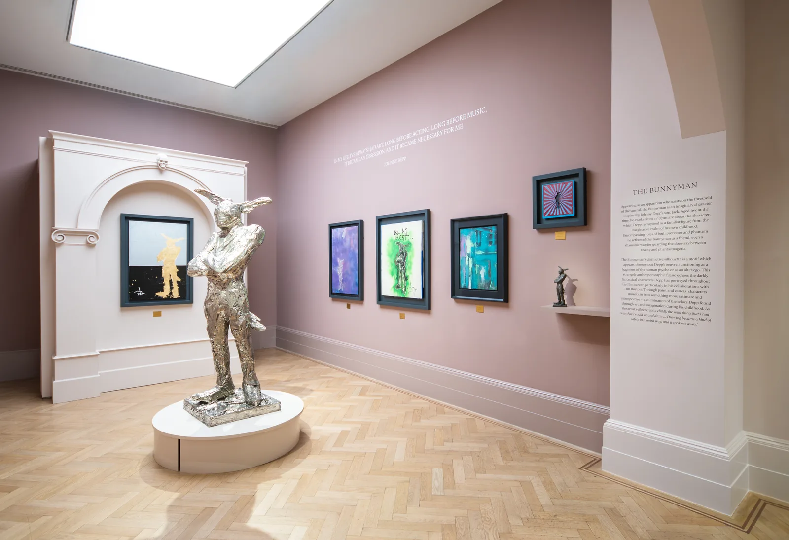 An overview of an ongoing solo gallery show highlights actor and artist Johnny Depp's debut major UK art exhibition, which opened on October 13, 2026, at London's Halcyon Gallery located at 148 New Bond Street. The display room is painted a soft dusty rose color, with a herringbone patterned wood parquet floor, a skylight overhead, and curated text printed directly onto the walls describing the artist's thematic concepts. Positioned on a low oval plinth in the left foreground stands a large textured white-bronze sculpture titled The Bunnyman, which depicts a two-meter tall half-human, half-rabbit guardian figure with folded arms. Behind it, framed within an ornamental classical arch built out from the wall, is a dark graphic artwork illuminated with gold leaf. Along the main back wall are three vertically aligned framed colorful paintings, accompanied by a smaller framed work with a striped background hanging higher up to the right and a miniature dark figurine sitting on a small ledge below it. This multimedia showcase spans decades of Depp's personal visual diary, tracking The Bunnyman character from its roots in his recurring childhood dreams and a drawing by his son Jack into various sketches, mixed-media paintings, and now his first-ever three-dimensional sculpture cast by Milan's Fonderia Artistica Battaglia.