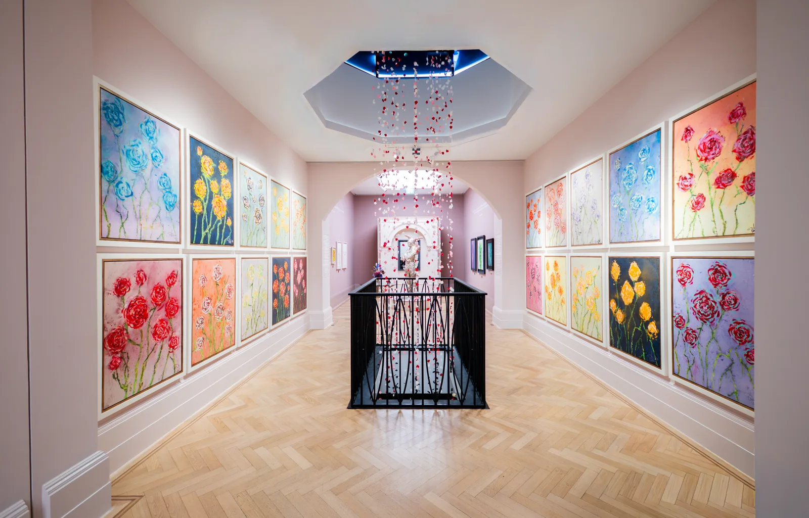 An elegantly lit, symmetric gallery hallway with light wooden herringbone floors and soft pink walls showcases a striking multi-piece exhibition of floral oil paintings on both sides. Framed in clean white borders, rows of colorful canvases depict heavily textured bouquets of yellow, periwinkle blue, and pink roses stretching toward an open archway at the far end. Suspended from an octagonal skylight in the ceiling, a whimsical, cascading string installation of red and white flower petals drops down over a black iron staircase railing in the center of the room. The vibrant, expressive paintings line the corridor leading toward a brightly illuminated sculpture niche, highlighting the expansive and deeply personal fine art showcase.