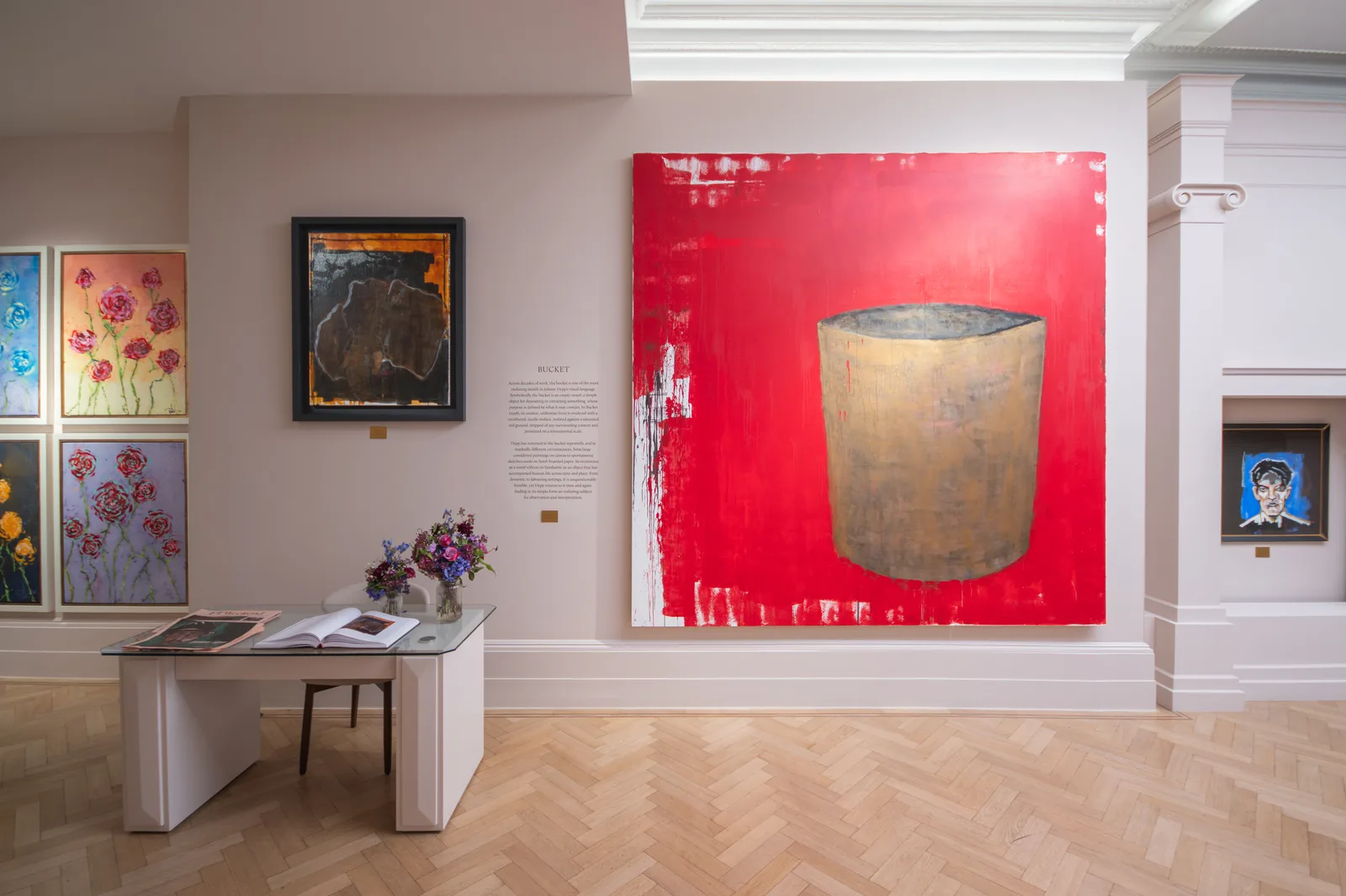 An exhibition gallery with light beige walls and a light wood herringbone parquet floor features several paintings by actor and artist Johnny Depp, anchored by a large, prominent canvas titled Bucket. The central, oversized artwork depicts a large tan or muted yellow bucket set against a vivid, heavily textured red background with white paint splatters on its left edge. To the left of Bucket, a framed text description explains the artwork, sitting above a small modern white writing desk with a glass top holding open gallery books and two small floral bouquets. Further left, a series of vertically stacked smaller paintings depicting floral motifs, including pink and deep red roses, line the wall. To the right of the central piece, an architectural white pillar frames another small, dark alcove showcasing a framed blue portrait.