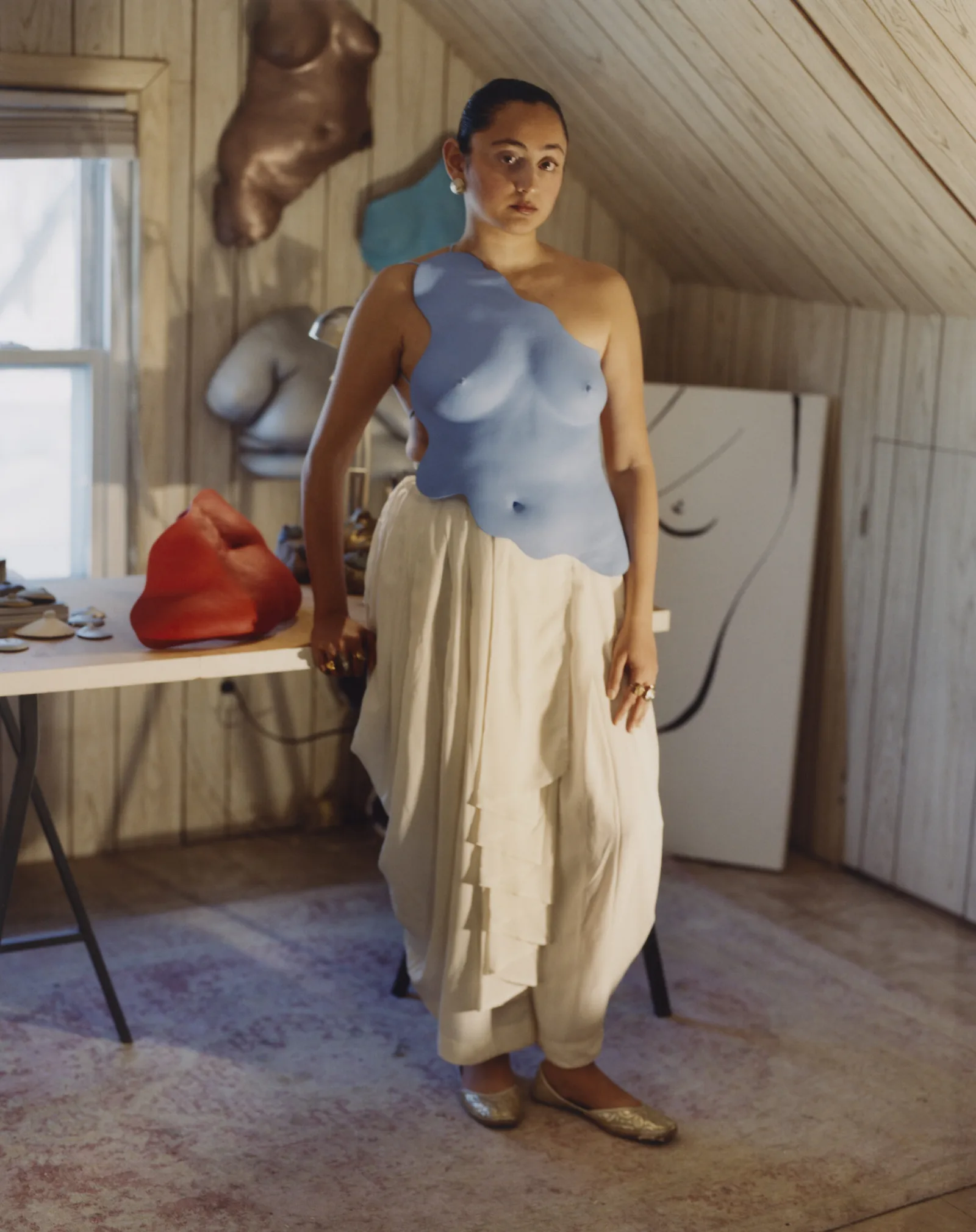 An elegant portrait of Pakistani visual artist and fashion designer Misha Japanwala standing inside a wooden-paneled attic studio. She is captured looking directly at the camera, wearing an asymmetric, one-shoulder sky blue sculptural breastplate molded directly from a female torso. She styles the statement armor over voluminous, draped white pants resembling a traditional dhoti or lungi, paired with gold metallic traditional flats. Her dark hair is slicked back into a neat bun, highlighting chunky gold statement earrings and a matching large ring on her finger. The artist's workspace is cluttered with creative materials, featuring several other multi-colored plaster anatomical casts—including a prominent red abstract form on a white worktable to her left, a metallic brown torso mounted on the back wall, and a large black-and-white minimalist outline drawing of a figure resting against the wall behind her. Soft, natural light streams in from a window on the left, casting subtle shadows across the splattered studio floor and emphasizing the tactile, raw qualities of both her wearable sculptures and her working environment.