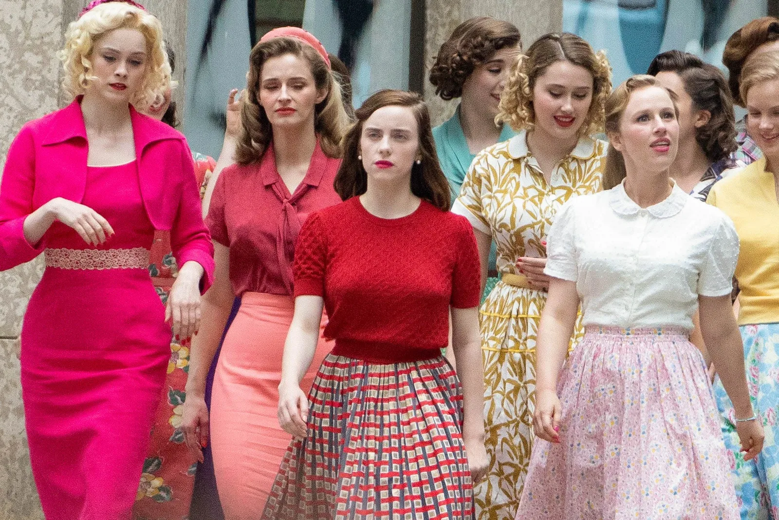 In this candid photograph taken on a bustling city street, a crowd of women are dressed in vibrant, mid-century 1950s attire. Walking in the center is the prominent figure of Billie Eilish, looking entirely unrecognizable and straying from her signature streetwear look. She is captured in character wearing a bright, form-fitting, cherry red short-sleeved knit top paired with a voluminous, pleated red-and-blue grid-patterned circle skirt. Her auburn-brown hair is meticulously styled into vintage, pinned curls, complemented by clean makeup, pearl earrings, and bold red lipstick. Surrounding her are background actors clad in complementary period costumes, including a woman to her left in a striking magenta dress and cropped blazer, and others in pastel floral dresses and tailored blouses. The setting mimics a 1950s New York City street during the filming of the movie adaptation of Sylvia Plath's classic 1963 novel, The Bell Jar, which marks Eilish's feature film acting debut.