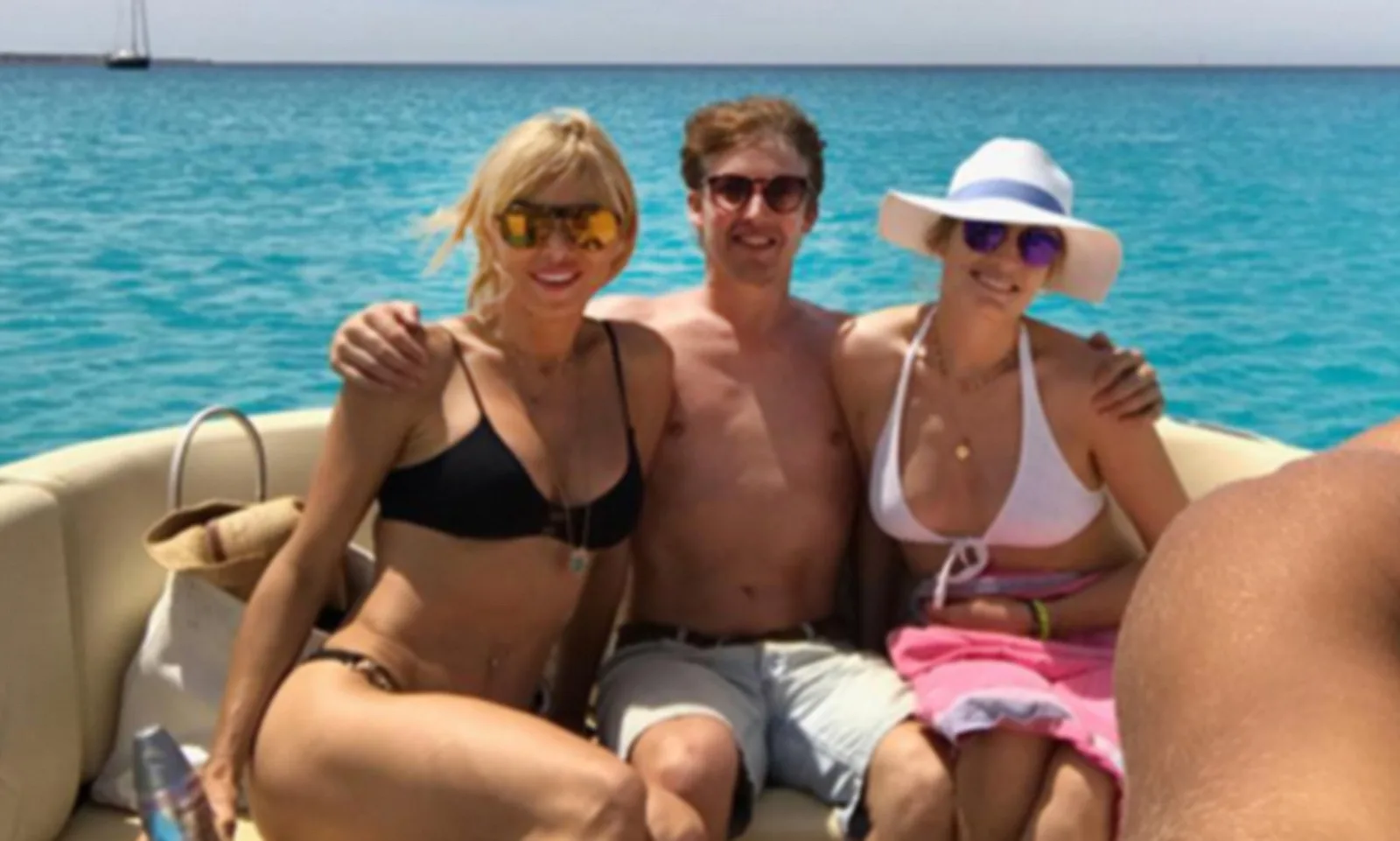 English singer-songwriter James Blunt is pictured shirtless on a cream-colored speed boat in Ibiza alongside two women, wearing sunglasses and enjoying a sunny summer day out on the ocean. Contrary to online speculation and rumors misidentifying the woman on the left as American actress Hayden Panettiere, the woman on the left in the black bikini is actually Bosnian entrepreneur and former Real Housewives of Beverly Hills star Sanela Diana Jenkins. The woman on the right smiling in a white halter bikini top, pink cover-up wrap, and a wide-brimmed sun hat is James Blunt’s wife, British socialite and legal consultant Sofia Wellesley.
