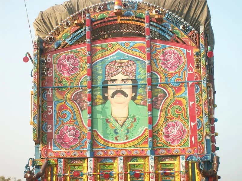 A detailed close-up shot showcasing the back of a Pakistani cargo truck adorned with vibrant, traditional truck art. The central focus is a large, hand-painted portrait of a man wearing a traditional Sindhi cap, possessing a thick black mustache, and dressed in a green shirt. The portrait is framed within an ornate, multicolored archway decorated with elaborate floral motifs, featuring large pink and white roses tucked into the corners amidst intricate swirls of yellow, blue, red, and orange paint. Flanking both sides of the central frame are vertical wooden panels painted with numerical digits, with Arabic numerals on the left and Urdu numerals on the right. The upper structure of the truck features a curved wooden framework painted in matching bright colors, topped by a heavy brown canvas tarp that covers the cargo area, all set against a clear, pale blue sky background.