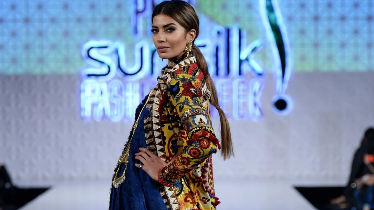 A Pakistani fashion model poses on the runway during a Sunsilk Fashion Week event, looking back over her shoulder toward the camera while modeling a striking multi-textured outfit. She wears a vibrant, heavily embroidered yellow and red jacket featuring intricate ethnic patterns, tassels, and geometric trim along the open lapels, layered over a deep royal blue draped dress or sari that features ornate gold coin embellishments down the front bodice. Her styled dark blonde hair is pulled back into a sleek, low, structured bubble ponytail that hangs over her shoulder, complementing her dramatic runway makeup look which includes defined brows, contoured cheekbones, and a matte nude lip. In the softly blurred background, the prominent, glowing blue and white neon signage for "Sunsilk Fashion Week" lights up the stage, framing her profile as she walks the white runway in front of a seated audience.
