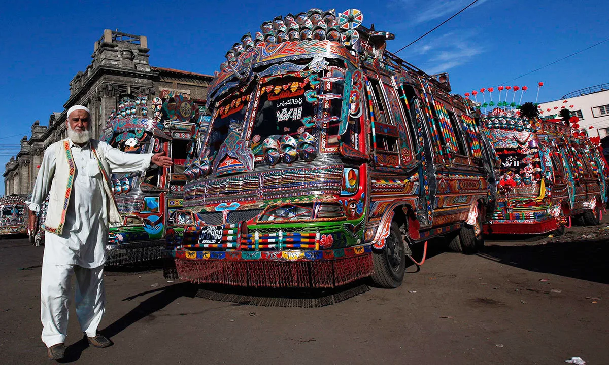 A wide outdoor shot under a clear blue sky captures a row of vibrantly decorated Pakistani mini-buses and trucks parked on a dirt lot, with a man standing prominently in the left foreground. The man, who has a long white beard and wears a traditional white shalwar kameez paired with a colorful embroidered vest, gestures with his left arm toward the vehicles. The main vehicle in the center is a highly customized bus covered from top to bottom in elaborate, multicolored truck art, featuring intricate geometric patterns, floral motifs, reflective ornaments, and a decorative fringe hanging below the front bumper. A license plate reading "JFO 460" is visible on its colorful front grille. To the left and right, other similarly ornate buses stretch into the background, parked in front of a weathered stone building with classical architectural details, while the dusty ground holds scattered debris.