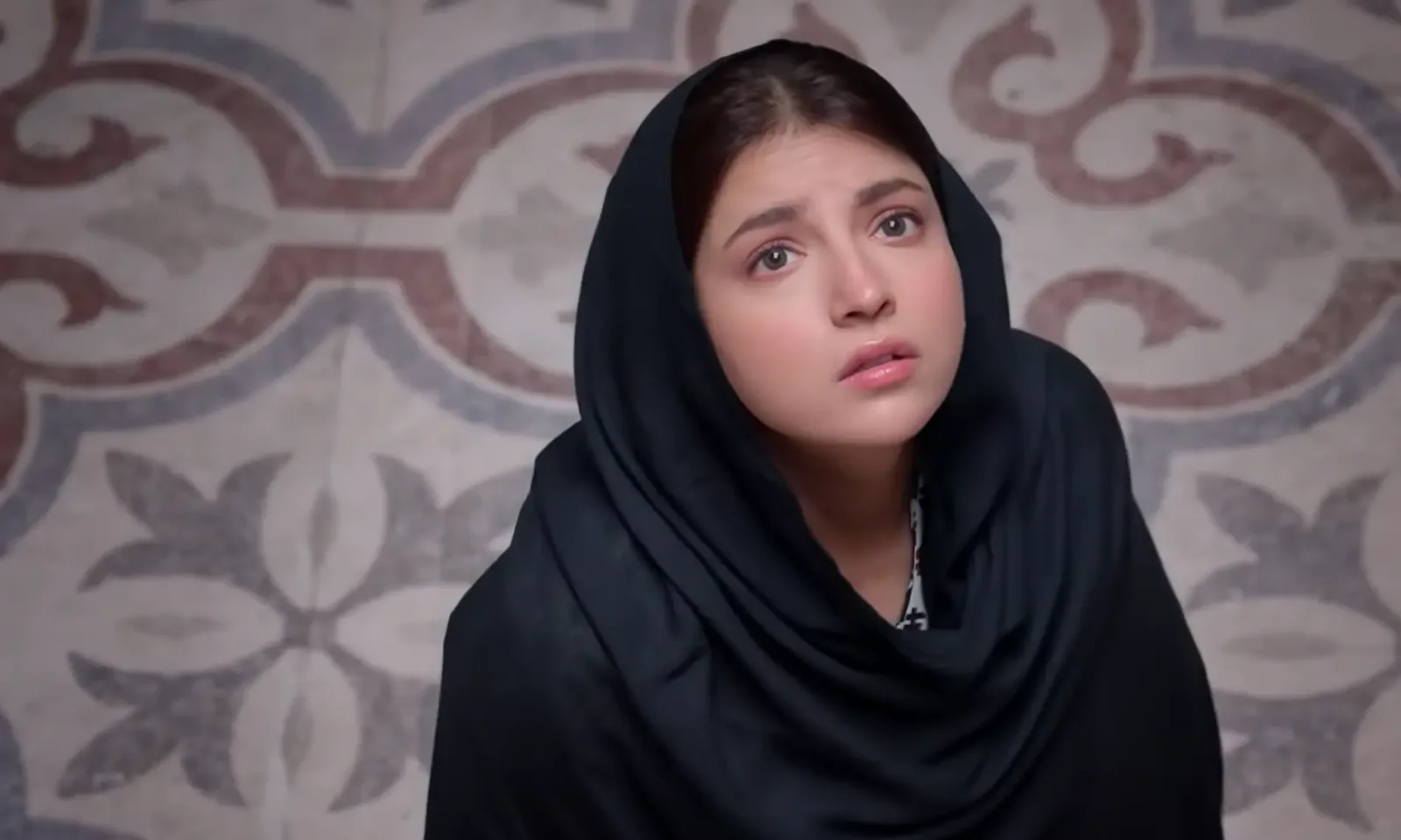 A close-up shot from the official teaser trailer of the television series showcases a young woman kneeling on a patterned floor, dressed in a plain black dupatta draped carefully over her head and upper body. Her expression is filled with an intense, vulnerable mix of longing and worry as she looks upward toward the sky, away from her immediate surroundings. Her fair complexion, soft makeup, and subtle tearful gaze emphasize the emotional weight of her character's situation. The detailed, classical tile patterns on the floor around her create a confined yet visually striking backdrop, reinforcing the themes of restriction, domestic confinement, and the yearning for personal freedom that define her emotional arc.
