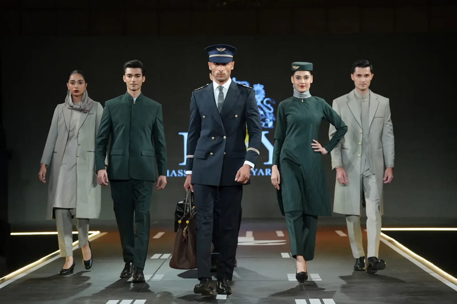 The image captures a striking group of fashion models confidently walking down a runway designed to mimic an airport tarmac, complete with white lane markings under warm, dramatic lighting. A male model dominates the center foreground, sharply dressed in a double-breasted navy blue pilot's uniform with gold button accents, a matching peaked cap, a crisp white shirt, and a dark tie, while carrying a structured brown leather duffel bag. To his right, a female model showcases a dark emerald green ensemble featuring a long tunic, matching trousers, a small green pillbox cap adorned with a gold emblem, and a light gray scarf neatly wrapped around her neck. On the left side, another female model wears a long, pale olive-drab trench coat over a textured cream-colored knee-length tunic and matching trousers, finished with a loosely draped taupe hijab. Flanking these central figures are two male models wearing sleek, modern outerwear: one in a minimalist teal-green mandarin-collar suit, and the other in a light beige overcoat layered over a matching buttoned jacket and trousers. In the soft-focus background, another male model stands wearing a bright neon-yellow security or safety vest over black attire, adding to the aviation atmosphere against a dark, minimalist backdrop bordered by vertical columns of golden light.