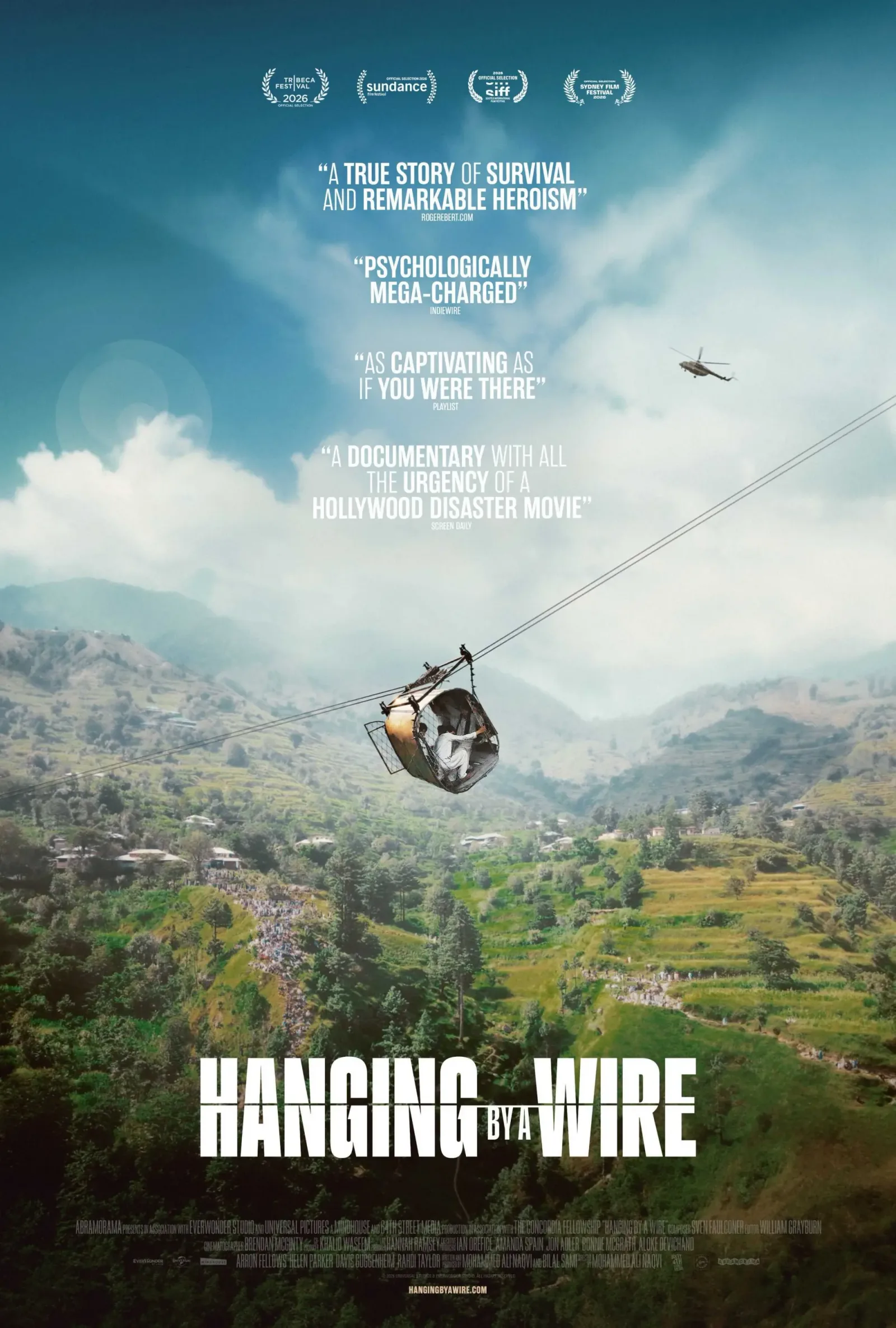 A vertical movie poster for the documentary "Hanging by a Wire" shows a small, open cable car carrying passengers high above a lush green valley in Pakistan. The cable car hangs precariously from a thin wire running diagonally across the frame, with steep, terraced hillsides, small village structures, and a crowd of onlookers visible far below in the background. In the upper right sky, a rescue helicopter approaches the scene under a bright, slightly hazy sky filled with soft clouds. Four positive critical review quotes are stacked vertically in white text above the cable car, topped by four film festival laurels, including Tribeca 2026 and Sundance. The film's title is boldly displayed across the lower third in a large, blocky white font, with billing block credits and a website URL printed in small text at the very bottom.