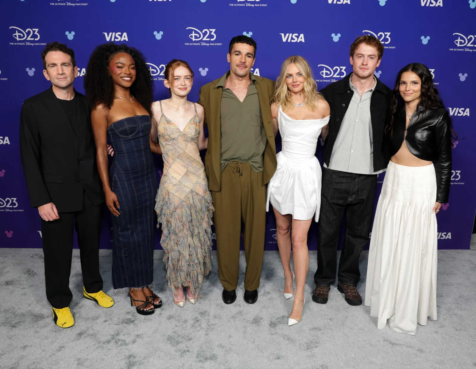 An ensemble cast poses together on a vibrant blue carpet featuring the D23 and Visa logos at a promotional entertainment event. From left to right, the group includes a man in an all-black suit paired with striking yellow sneakers; a woman with voluminous curly hair wearing a strapless, dark denim midi dress; a woman with red hair in a textured, strapless beige-and-white gown; a man in a relaxed olive-green suit over a matching button-down shirt; a woman with blonde hair wearing a draped, structured white mini dress with white heels; a man wearing a white collared shirt under a black jacket with dark trousers; and a woman in a black cropped leather jacket paired with a flowing, layered white maxi skirt. The actors stand closely in a line, smiling and posing for photographers against the branded media backdrop.