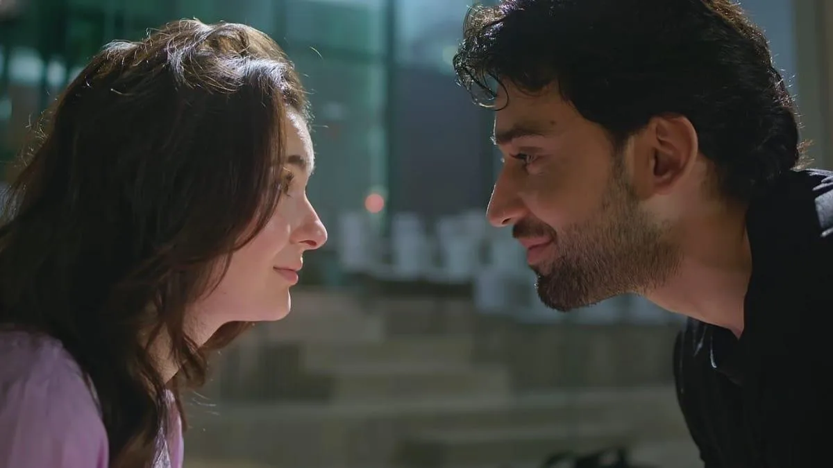 An intimate close-up shot captures the profound, unspoken chemistry between actors Hania Aamir and Bilal Abbas Khan as they lock eyes in a tender moment from a cinematic scene. Positioned in profile against a softly blurred, neutral-toned modern architectural backdrop, the pair are leaned in close to one another, their expressions radiating a deep and quiet affection. The woman on the left, with her dark hair falling softly around her shoulders and a gentle, serene smile playing on her lips, gazes up at the man. He returns her look with a warm, knowing smile, his dark, curly hair and light stubble framed perfectly by the dramatic lighting that highlights the contours of his face and dark shirt. The shallow depth of field isolates the duo completely, intensifying the romantic tension and emotional vulnerability shared between their characters in this beautifully composed frame.
