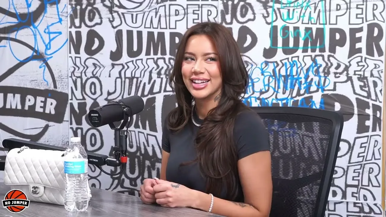 A medium close-up shot from a video interview features a young woman with long, voluminous brunette hair parted in the middle, smiling during a conversation. She is wearing a tight-fitting short-sleeved black t-shirt, a thin silver chain necklace, and a delicate beaded bracelet on her left wrist. She sits behind a grey wooden desk with her hands lightly resting on the surface. To her left, a professional black Shure microphone is mounted on an arm, positioned next to a clear plastic water bottle and a white quilted Chanel handbag. The background consists of a white wall densely covered in a repeating black-and-white graffiti-style "NO JUMPER" logo print.