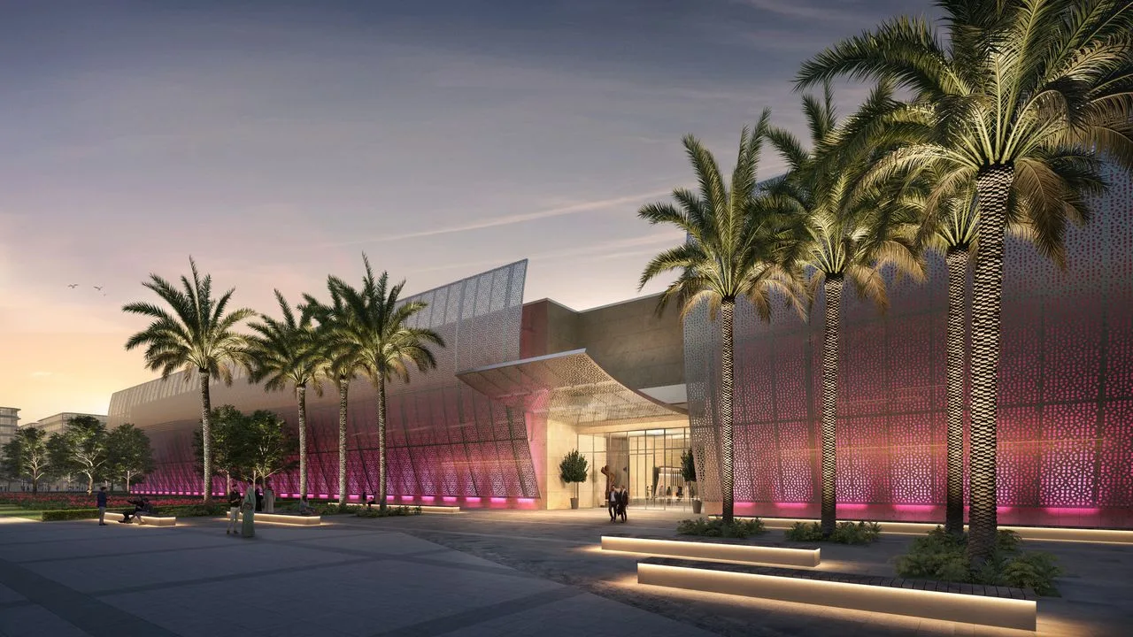 The image depicts the modern exterior facade of the Manarat Al Saadiyat cultural center, illuminated at dusk with vibrant pink and violet lighting filtering through an intricate mesh lattice overlay. Tall, well-manicured palm trees line the wide paved pedestrian plaza in front of the entrance, where integrated glowing light strips on the ground and stairs complement the contemporary architectural design. The rendering showcases the venue prepared for the highly anticipated inaugural Frieze Abu Dhabi art fair, a landmark collaboration with the Department of Culture and Tourism – Abu Dhabi that replaces the long-running Abu Dhabi Art platform.