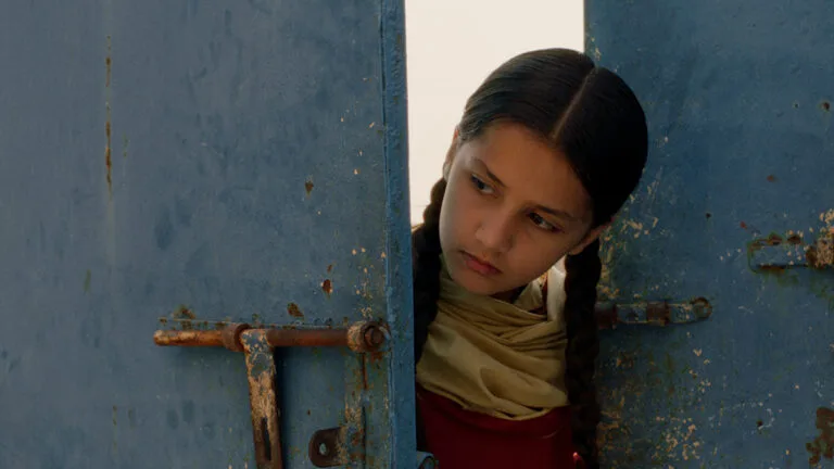 A still image from the Pakistani feature film Ghost School (2025) shows a young girl named Rabia (played by Nazualiya Arsalan) peering out from behind a distressed blue metal door. Her dark hair is parted down the center and tied into two braids that rest over her shoulders, and she wears a cream-colored scarf around her neck. She has a serious, curious expression as she looks to the side. The bright, overexposed backdrop contrasts sharply with the weathered, peeling blue paint of the double doors, which feature a rusted sliding iron latch on the left panel. The composition frames her head in the narrow opening between the two heavy doors, creating a sense of hidden observation.