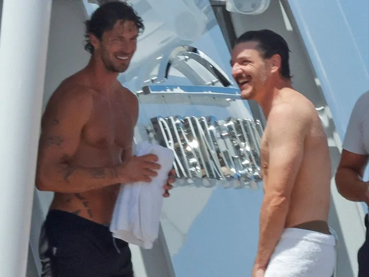 Actor Pedro Pascal stands alongside fashion model Christian Hogue on the deck of a sunlit superyacht, both appearing shirtless and wrapped in white towels. The candid snapshot captures the pair mid-conversation, sharing a genuine laugh during a star-studded Mediterranean getaway that also included celebrity friends like Jason Bateman and Judd Apatow.