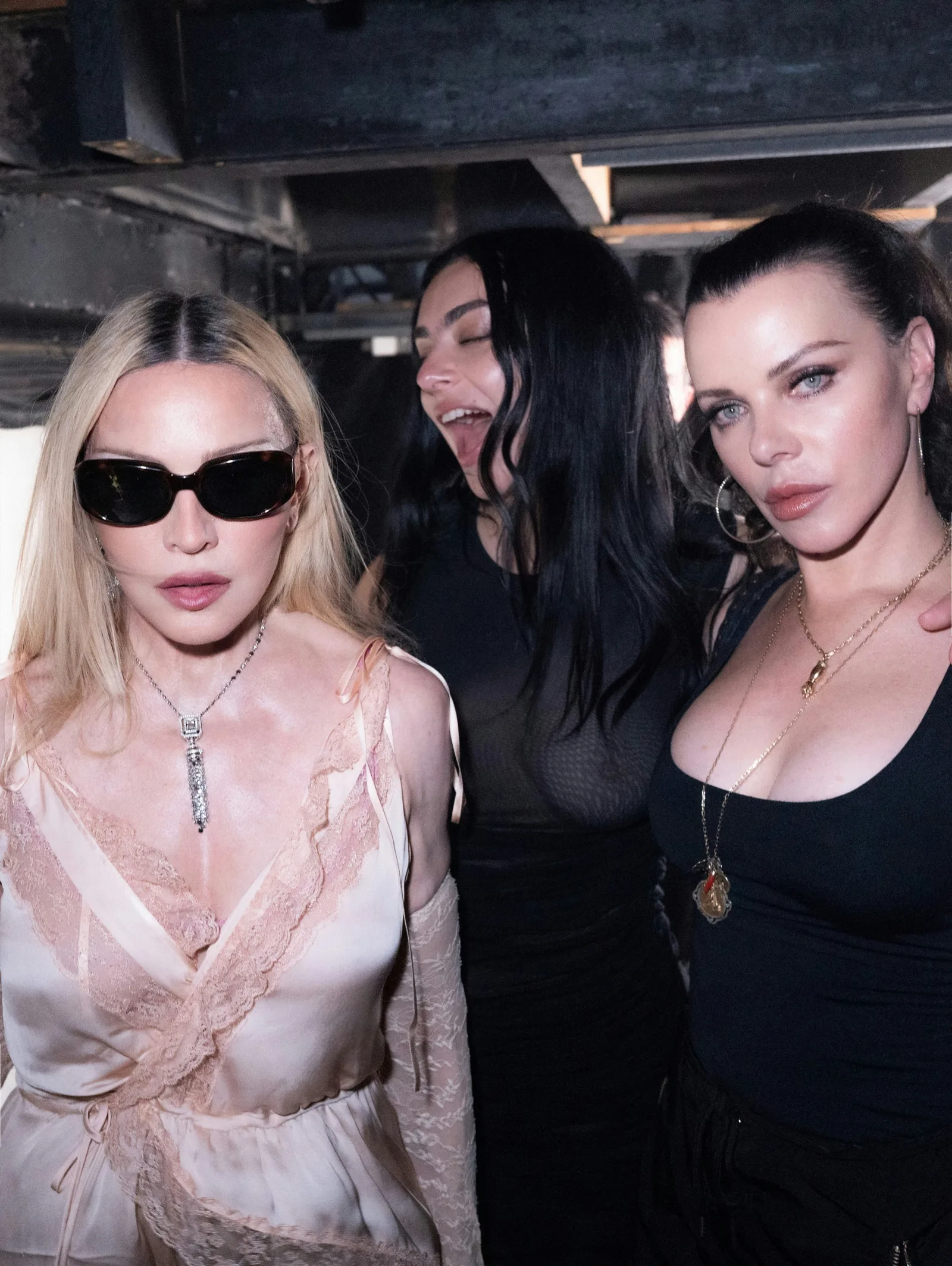This candid shot captures the iconic Madonna, British pop sensation Charli XCX, and actress Julia Fox sharing an intimate, high-energy moment together during an exclusive late-night gathering. Madonna leads the frame on the left, exuding timeless cool in a silk-and-lace cream slip dress paired with stark black sunglasses and a statement pendant necklace. Beside her, Charli XCX is caught mid-laugh with her eyes closed, radiating carefree energy in a sheer black top that adds a moody, electronic-pop edge to the scene. Grounding the right side of the composition is Julia Fox, who fixes the camera with an intense, smoldering gaze, wearing a minimalist black scoop-neck tank top complemented by layered gold chains and classic oversized silver hoop earrings. The raw, low-ceilinged industrial background and sharp direct flash create a gritty, backstage party atmosphere, perfectly encapsulating a powerhouse meeting of distinct generation-defining pop culture and fashion figures.