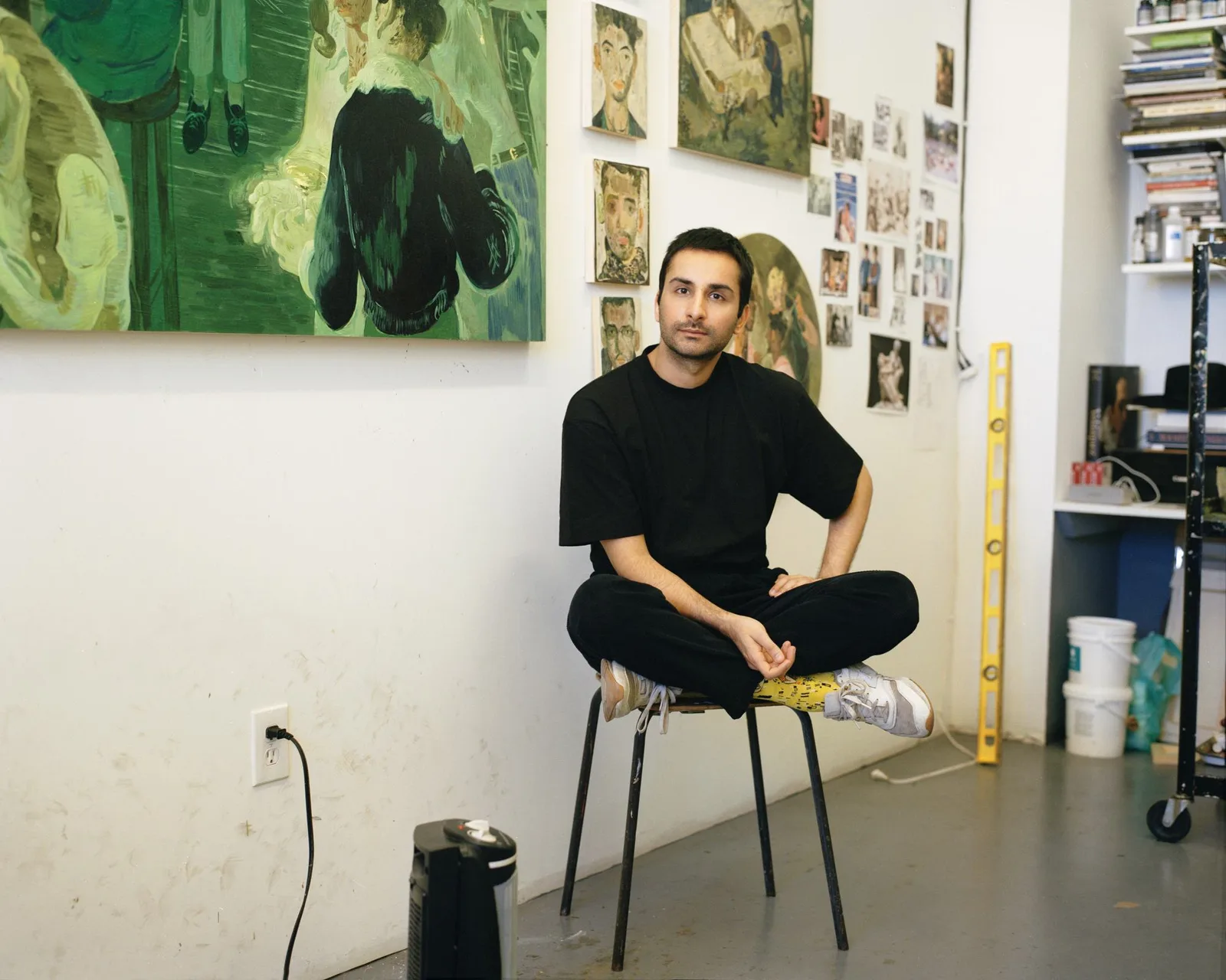 Salman Toor sits cross-legged on a metal-legged stool in the center of his brightly lit art studio, looking directly at the camera with a calm expression. He is wearing a loose-fitting black crewneck T-shirt, black trousers, patterned socks, and grey sneakers. Behind him, a stark white wall is adorned with various works of art, including a large green-hued painting on the upper left depicting figures, smaller painted portraits, sketches, and reference photographs clustered together. To his right, a tall yellow spirit level rests against the wall near a white bucket, a desk piled with books, and the edge of a rolling cart, while an electric humdifier sits on the floor to his left next to a wall outlet.