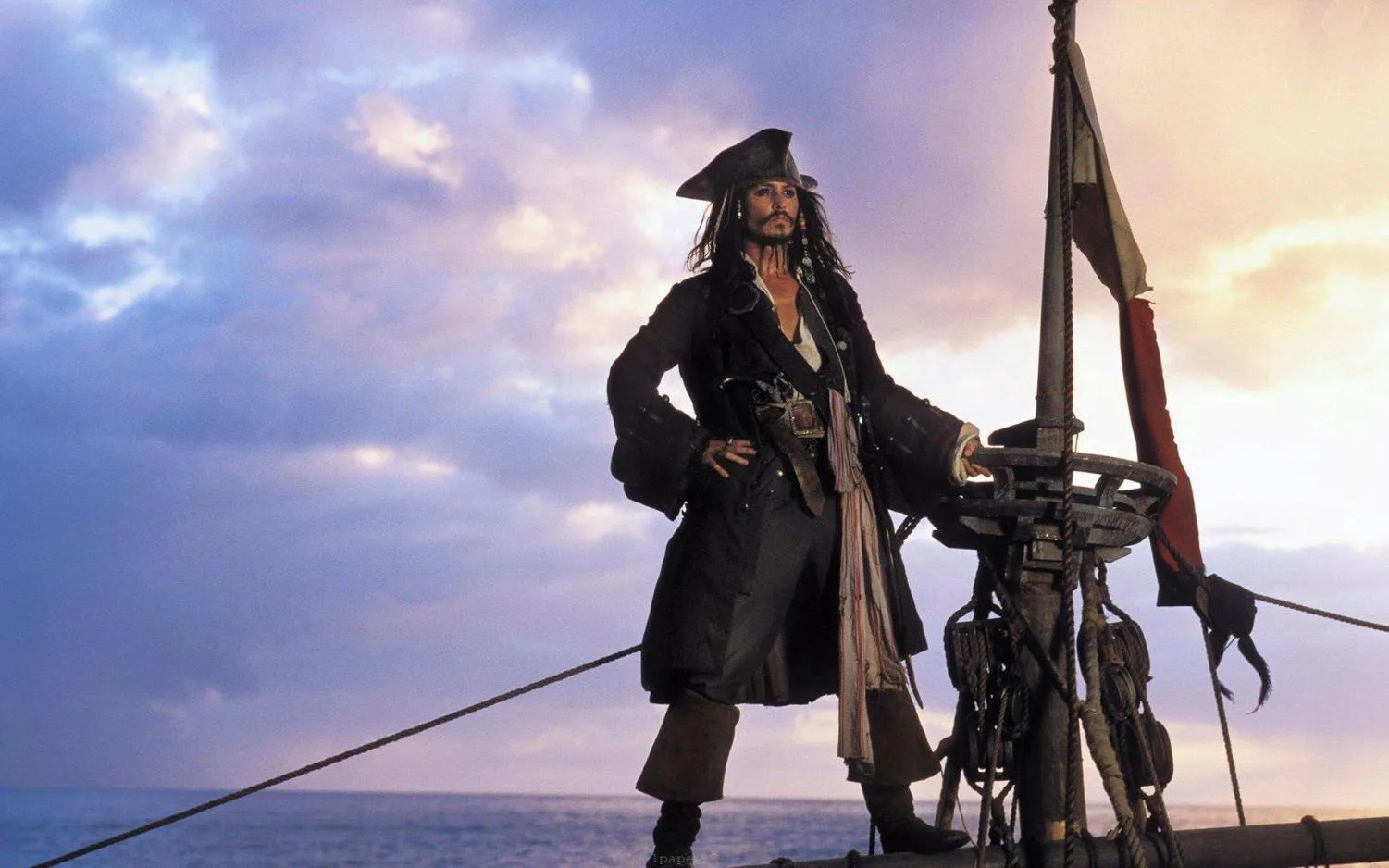An iconic medium shot captures Captain Jack Sparrow standing proudly at the bow of a wooden pirate ship under a vast, dramatic sky filled with soft lavender and gold-tinted clouds. He is dressed in his signature pirate attire, including a dark tricorn hat, a long dark frock coat over a vest, a wrapped fabric sash around his waist, and heavy knee-high boots. Standing with a confident stance, one hand rests on his hip while the other grips the ship's rigging or structure near a small tattered flag. The background reveals a wide, open ocean extending to the horizon under the lighting of either dawn or dusk.