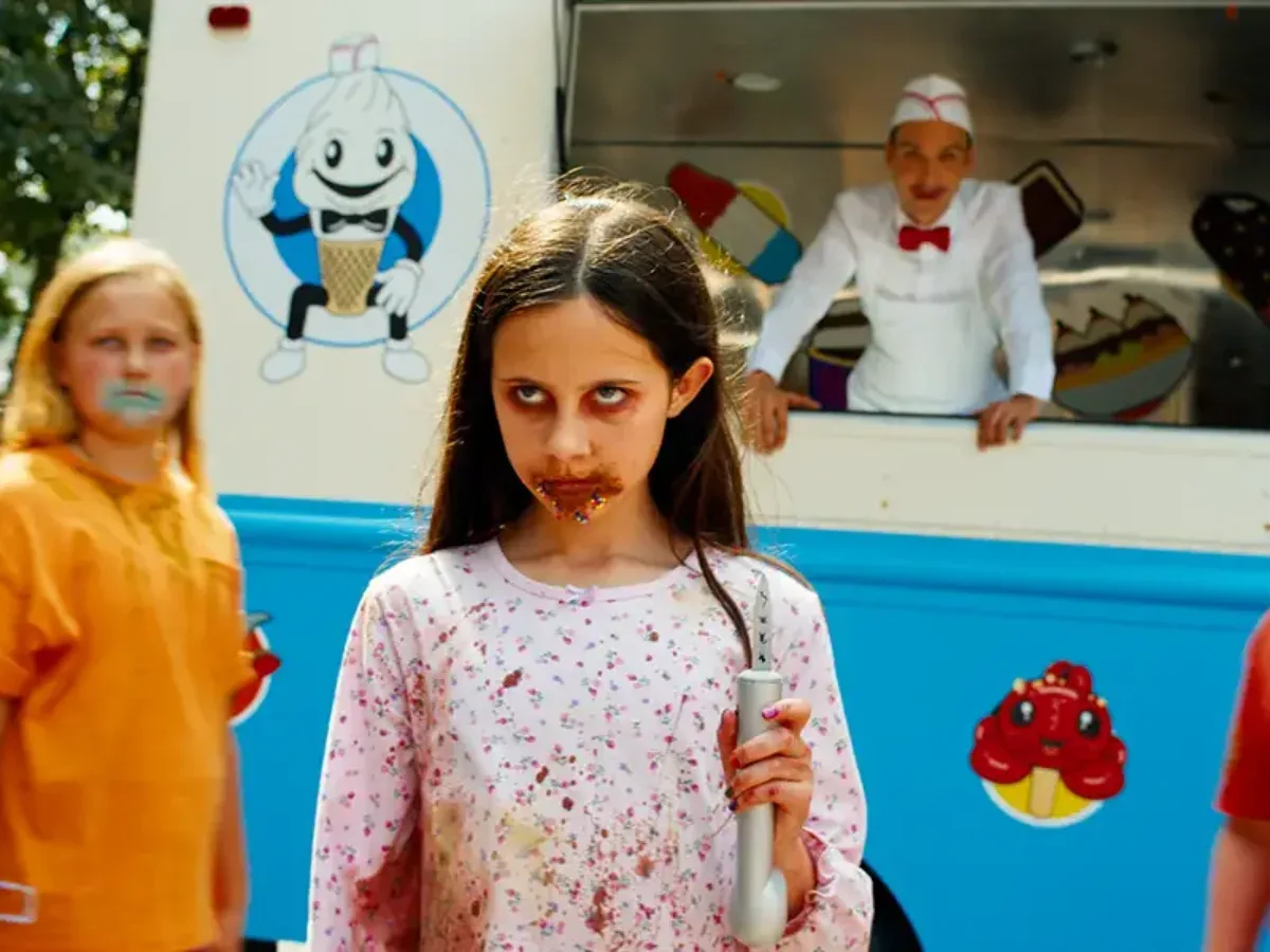 A high-intensity, sunlit movie still capturing a deranged, dark-haired young girl standing menacingly in the foreground, staring intensely ahead with hollowed, dark-circled eyes and a mouth smeared with a gritty, brown chocolate mixture that resembles dried blood and sprinkles. She wears splattered pink floral pajamas and holds a silver object in her hand. Behind her to the left, another young girl in an orange shirt watches blankly with blue popsicle staining her lips, while an eerie ice cream man dressed in a crisp white uniform, matching paper hat, and a red bow tie leers forward with an unsettling smile from the window of a bright blue and white ice cream truck decorated with a cartoon ice cream cone mascot.