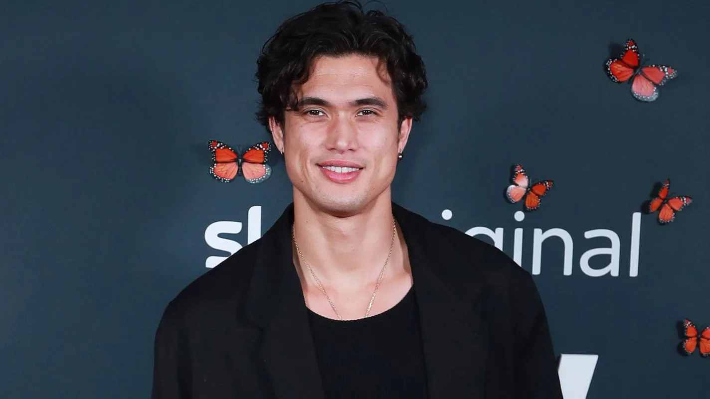 Charles Melton - An attractive young man with short, wavy dark brown hair and a warm smile stands against a dark grey backdrop decorated with several scattered orange monarch butterflies. He wears a simple black scoop-neck shirt layered underneath an oversized black blazer, complemented by a delicate, thin gold chain necklace.