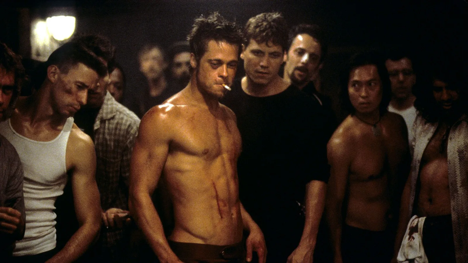 A shirtless, lean, and muscular Tyler Durden (Brad Pitt) stands at the center of an underground fight club basement, a lit cigarette dangling from his mouth and faint bloody cuts visible on his torso. He is surrounded by a dense, intense crowd of onlookers and fighters, including a man in a white tank top on the left and several other men staring intently from the shadows. The dimly lit environment captures the gritty, raw atmosphere of David Fincher's iconic psychological drama.
