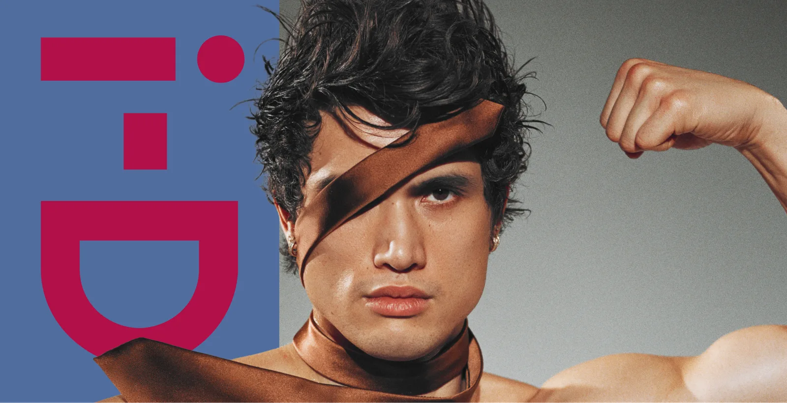 A close-up studio photograph features actor Charles Melton for i-D Magazine, sporting dark, wavy, textured hair and a light stubble. A wide, glossy satin ribbon in a rich bronze-brown hue wraps dramatically around his head, covering his right eye like a makeshift blindfold before draping down around his neck. He poses with his left arm raised and flexed, showing off his bicep against a neutral gray background. On the left side of the frame, a solid blue vertical block features the bold, pink geometric lettering of the "i-D" magazine logo.
