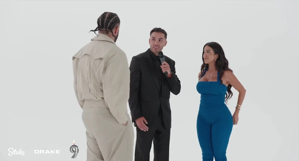 In a brightly lit, sterile white studio environment, Canadian rapper Drake stands with his back partially turned to the camera on the left, sporting braided hair and wearing an oversized, tan, utilitarian-style long-sleeve shirt paired with matching khaki trousers. In the center, Kyle Forgeard of the Nelk Boys looks toward Drake while speaking into a handheld black microphone, dressed in a sharp, monochromatic black suit and black button-down shirt. On the right, internet personality Lena the Plug stands facing the men with a slight smile, styled in a vibrant, form-fitting blue halter-neck jumpsuit with her long, wavy brunette hair flowing down her shoulders.