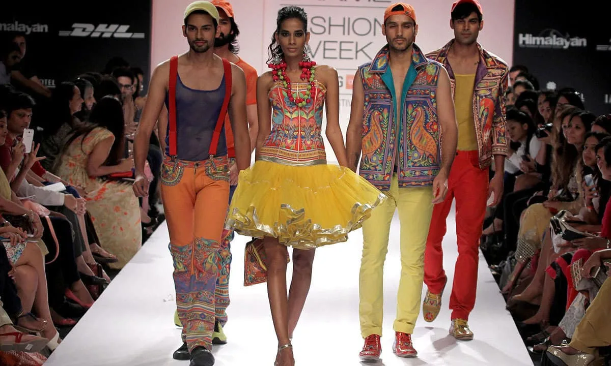 A vibrant runway display from Lakme Fashion Week showcases a contemporary collection deeply inspired by South Asian truck art, featuring models in highly stylized, kaleidoscopic garments. The outfits burst with saturated hues of bright yellow, vivid orange, and electric blue, meticulously embellished with intricate, multicolored patterns, peacock motifs, and ornate borders reminiscent of traditional transport decoration. The ensemble blends street-style silhouettes like structured vests, suspenders, and layered mesh tops with dramatic elements, including a voluminous yellow tutu-style tiered skirt, accessorized with heavy beaded necklaces and playful forward-facing caps. A vibrant fashion runway show features four models walking side by side down a white catwalk flanked by a seated audience. On the far left, a male model wears a sheer blue tank top with red suspenders, light orange trousers featuring patterned panels at the lower legs, and a lime green cap. Next to him, a female model walks confidently in a colorful patterned strapless corset top paired with a voluminous, layered bright yellow tutu skirt featuring metallic silver trim along the hemline. To her right, another male model showcases an open, sleeveless vest adorned with intricate orange and blue peacock motifs over a teal lining, paired with pale yellow pants and an orange cap. On the far right, a male model walks in a yellow tank top beneath an elaborately patterned multi-colored jacket, matching red trousers, and a black cap. The background features a large event backdrop displaying "FASHION WEEK" along with corporate sponsor logos like DHL and Himalayan, under soft, dramatic runway lighting.