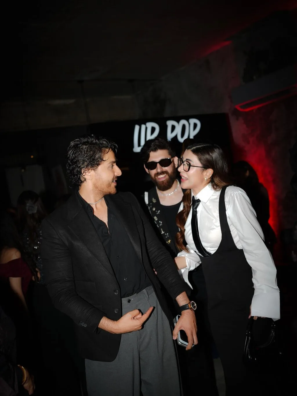 Urwa Hocane and Ahsan Khan at the Sunday Icon Awards Nominees Reveal Party. An image capturing a candid moment of three individuals at an indoor nighttime gathering, likely an event or party. The person on the left is a man seen in profile, facing right. He has wavy, dark hair, a light mustache and beard, and is wearing a black blazer over a black button-down shirt paired with grey trousers. His left arm is extended downward, holding a smartphone, while his right hand points toward his waist area. The individual on the right is a woman with long, dark brown hair, wearing thick-rimmed glasses. She is dressed in a white button-down dress shirt with an attached slim black tie, styled under black overall-style trousers. She is facing left toward the man, smiling as they appear to be in conversation. Positioned slightly behind and between them is another man with a full beard, wearing dark sunglasses, a black top, and a necklace. The background is dimly lit with a dark ambiance, featuring red accent lighting on the right wall and a blurred sign in the upper-center that reads "LIP POP." Other party attendees are faintly visible in the dark background.