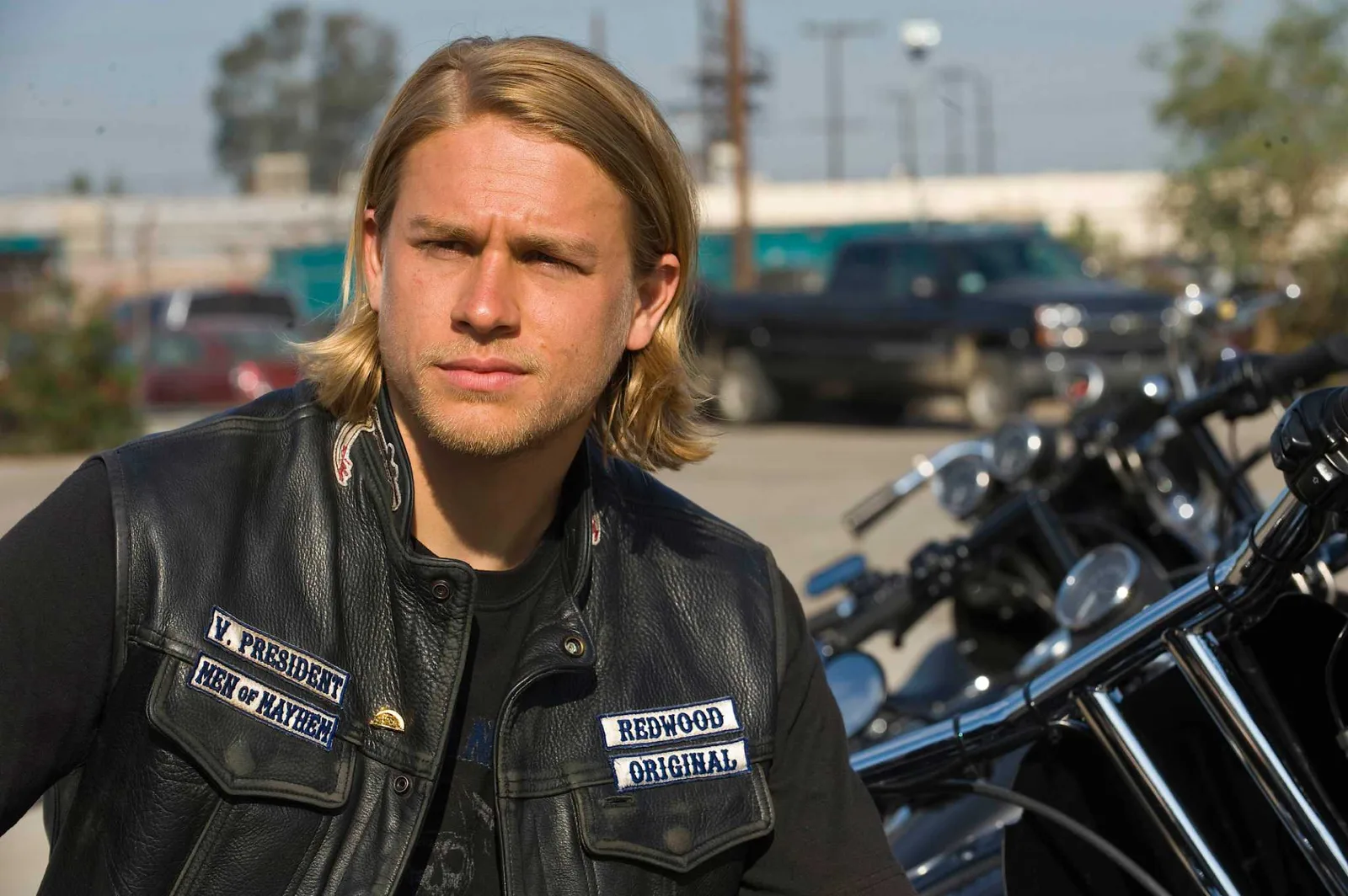 The image features actor Charlie Hunnam starring as the protagonist Jax Teller in the acclaimed crime drama television series Sons of Anarchy. He is depicted with his signature shoulder-length blonde hair and light stubble, wearing a black leather motorcycle club vest adorned with official patches including "V. PRESIDENT", "MEN OF MAYHEM", and "REDWOOD ORIGINAL" over a casual black t-shirt. He stands outdoors in a sunny, industrial-style lot with a line of heavy cruiser motorcycles parked closely in the foreground and a dark pickup truck visible in the soft-focus background, capturing the gritty, rebellious essence of the show's outlaw motorcycle club culture.