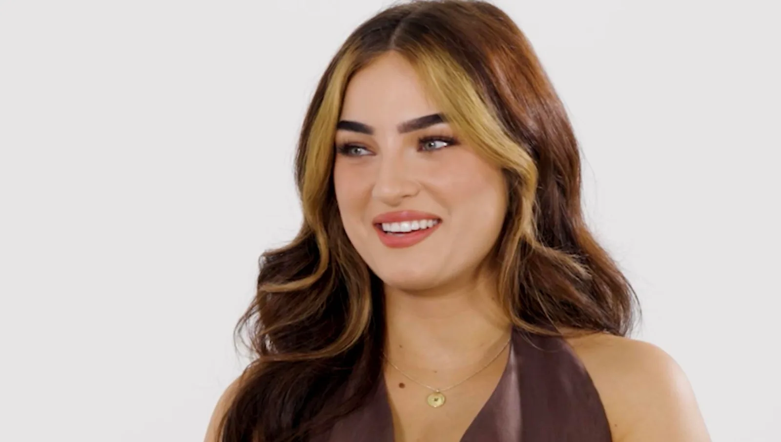 A medium close-up shot from an interview showing filmmaker Savanah Moss seated on a dark gray or black tufted leather chesterfield sofa against a solid white background. She is turned slightly to her right, looking off-camera with a friendly expression and gesturing with her hands while speaking. She has long, dark brown hair styled in soft waves with prominent blonde face-framing highlights. She wears a brown button-up sleeveless vest matching her trousers, a delicate gold pendant necklace, a gold bangle on her right wrist, and features manicured white nails. A small, white "Forbes" logo is visible in the bottom right corner of the frame. Savanah Moss & Ian Boggs Talk Brand Deals And Favorite Partnerships. This interview screenshot features digital creator and film director Savanah Moss speaking about her creative career and upcoming projects.