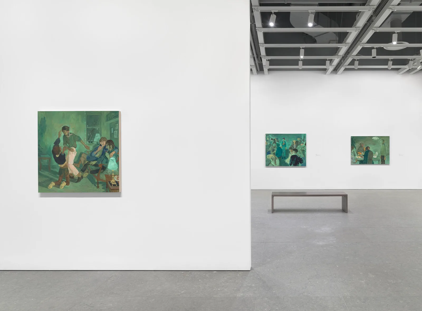 An installation view captures the solo exhibition Salman Toor: How Will I Know at the Whitney Museum of American Art in New York. Set within a spacious, white-walled gallery under an exposed industrial ceiling grid equipped with gallery track lighting, the photograph highlights three figurative oil paintings that showcase the artist's signature green-tinged color palette. On the left, mounted prominently on a dividing wall, is the piece Four Friends (2019), depicting an intimate interior scene where stylized, long-limbed young men engage with one another in a warm, domestic setting. Further down the gallery floor, a simple dark wood minimalist bench sits parallel to the back wall, where the remaining two artworks are hung side-by-side: Bar Boy (2019) in the center—which features a central figure illuminated by the glow of a smartphone screen among others—and Tea (2020) on the right. The combination of polished gray concrete flooring, extensive white wall space, and deep emerald and sage canvases underscores Toor's focus on vulnerability, intimacy, and contemporary queer community spaces.