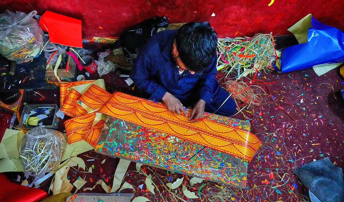 An overhead shot captures a male artisan sitting cross-legged on a dark floor in a cluttered workspace, meticulously crafting a long banner of vibrant orange truck art paper adorned with detailed yellow and white patterns. He is wearing a dark blue long-sleeved shirt and trousers, focusing closely as his hands manipulate the material over a worn wooden workbench covered in a colorful explosion of tiny paper scraps. The surrounding environment is densely packed with tools and materials, including plastic bags filled with cut paper strips, large sheets of red and bright blue paper, scattered shavings, and trimmings on the floor, all framed against a rough, textured red wall at the top of the frame.