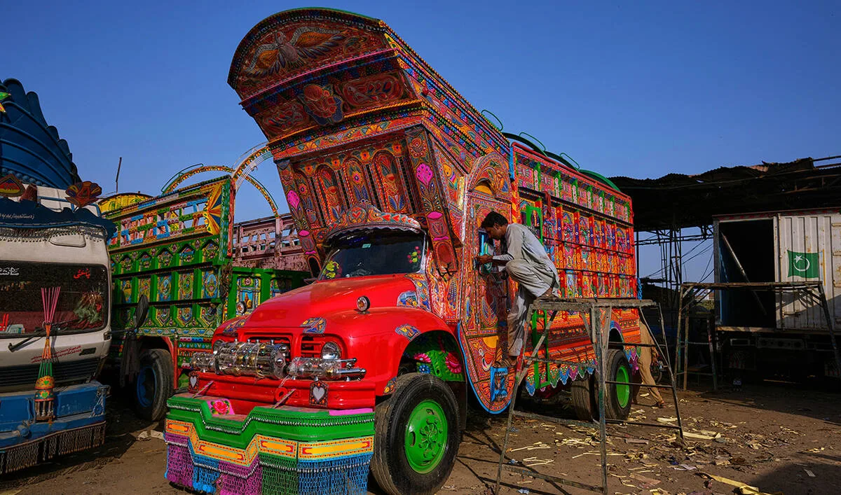 A brightly painted cargo truck features a vibrant red hood and an towering, ornate wooden crown extending far above the cab, completely covered in intricate geometric patterns, floral motifs, and bold pink, orange, and blue hues. A craftsman stands precariously on a metal ladder propped against the driver's side door, meticulously painting fine details onto the vehicle's heavily decorated exterior within a bustling outdoor workshop.