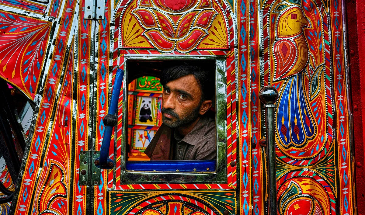 A man with dark hair and a beard peers out from the rectangular window of a highly decorated vehicle, surrounded by the intricate, vibrant patterns of Pakistani truck art. The exterior paneling bursts with bold hues of red, orange, yellow, and blue, featuring geometric borders, floral motifs, and a stylized depiction of a bird on the right side. Through the small window opening, the vehicle's interior reveals further artistic details, including small painted panels of a panda and a parrot alongside Urdu script, creating a visually dense and culturally rich composition.