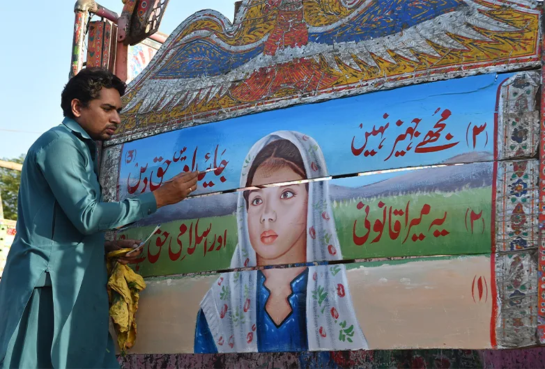 A detailed photograph captures a Pakistani truck artist meticulously painting a large, vibrant mural on the wooden back panel of a cargo truck. On the left, the artist, a man with dark hair, a mustache, and a focused expression, wears a traditional teal shalwar kameez while holding a thin paintbrush in his right hand and a yellow cleaning cloth in his left. The centerpiece of the mural is a realistic portrait of a young girl with large, expressive eyes, wearing a white dupatta over her head with blue and red floral patterns, and a blue shirt. The background of her portrait features rolling green hills under a soft blue and purple sky. Surrounding the portrait are multiple panels filled with elaborate Urdu calligraphy written in bold red and white scripts against blue, green, and white backgrounds, outlined with delicate floral borders. Above the central artwork, a large, stylized wooden carving of a bird with widespread wings is decorated with intricate, multi-colored geometric and floral patterns in shades of yellow, red, blue, and white, showcasing the rich tradition of Pakistani truck art.
