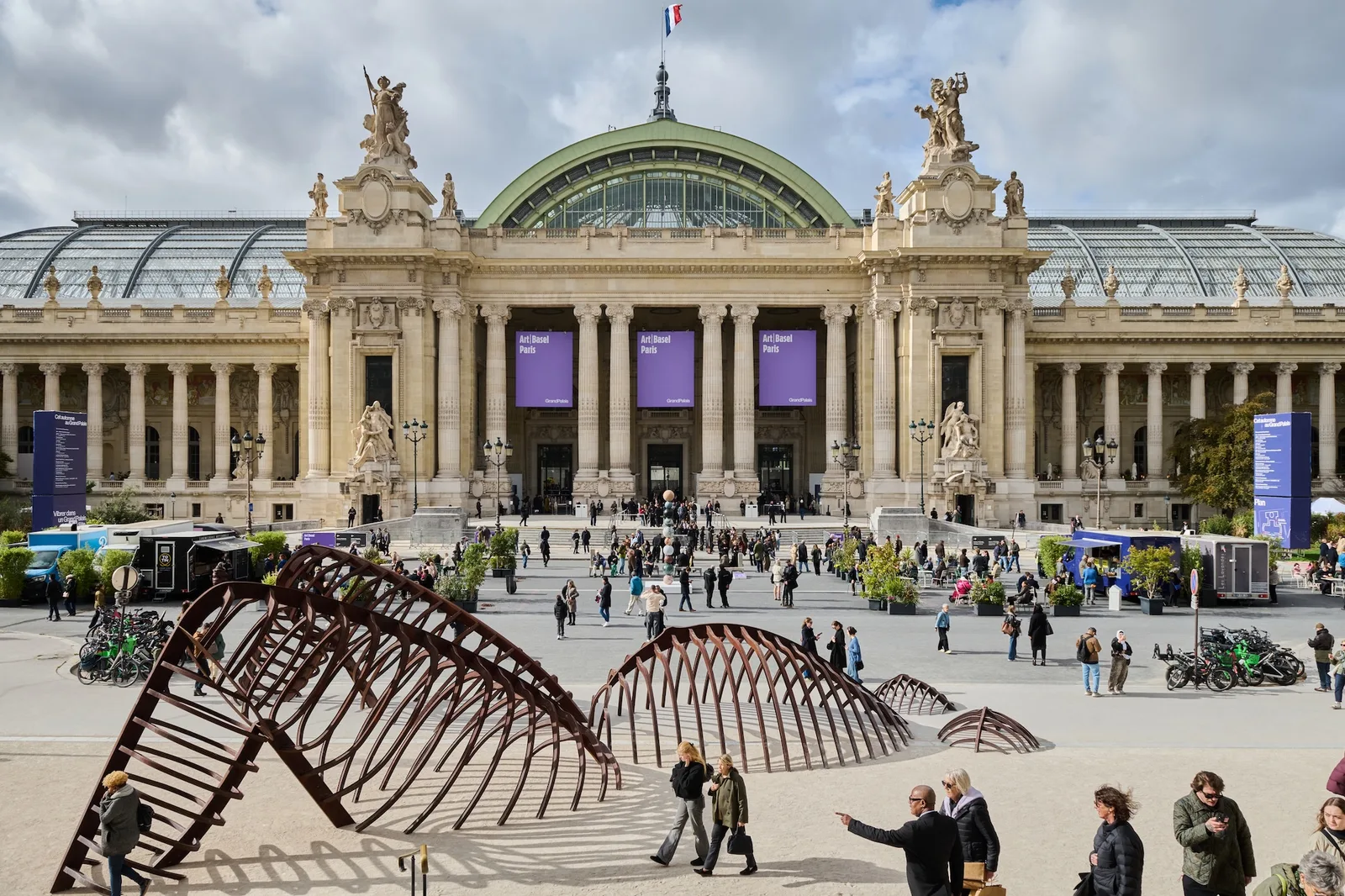 An outdoor scene captured on Avenue Winston Churchill in Paris showcases monumental art installations displayed as part of the public program for Art Basel Paris, positioned right in front of the ornate, golden-gated entrance of the historic Petit Palais. On the left stands L'Élévation des Mondes (2026) by artist Alicja Kwade, a striking vertical tower consisting of stacked, multi-colored stone orbs balancing on a white rectangular pedestal. To its right, a series of dark, arched, rib-like metal structures curve over the pedestrianized street, drawing the attention of scattered onlookers who wander through the exhibition space. In the foreground, concrete geometric benches invite visitors to sit, with one individual viewed from behind resting on a bench, while blurred figures of pedestrians evoke the constant motion of the city around the static public sculptures. Large exhibition banners featuring classical portraits hang from the classical stone facade of the palace building under a bright, overcast sky.