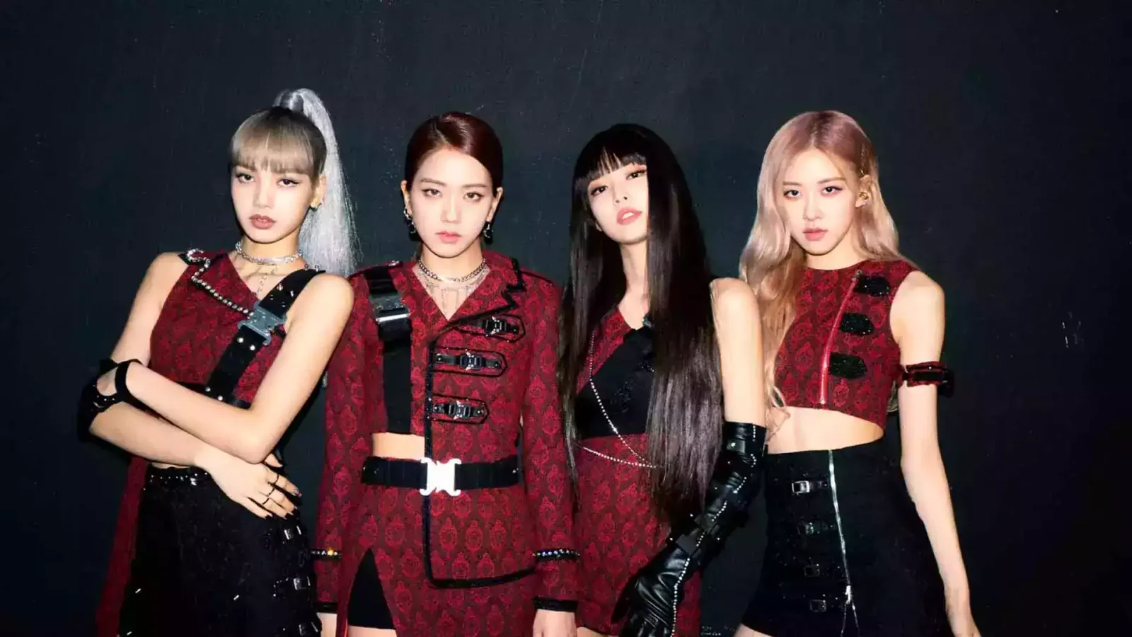 An elegant group portrait captures the four members of the South Korean girl group BLACKPINK standing side-by-side against a stark, solid black background. From left to right, Lisa, Jisoo, Jennie, and Rosé pose confidently in coordinating, high-fashion stage outfits dominated by deep crimson brocade patterns and edgy black accents. Lisa sports a high, sleek platinum-blonde ponytail with blunt bangs, wearing a sleeveless patterned top crossed by black tactical buckled straps. Beside her, Jisoo wears her dark hair parted to the side, styled in a tailored, structured crimson blazer-style dress featuring prominent metallic buckles and a thick black belt at the waist. Jennie leans slightly back with a fierce expression, her long, straight black hair framing her face, dressed in a matching patterned top paired with dramatic, shiny black patent leather opera gloves. On the far right, Rosé showcases long, wavy rose-gold hair, wearing a sleeveless, cropped crimson top adorned with black button details that exposes her midriff, paired with a high-waisted black skirt featuring a prominent silver zipper running down the front. The sharp, direct studio lighting casts soft shadows behind them, highlighting the intricate textures of their dark, rebellious-chic ensembles and their intense, captivating expressions.